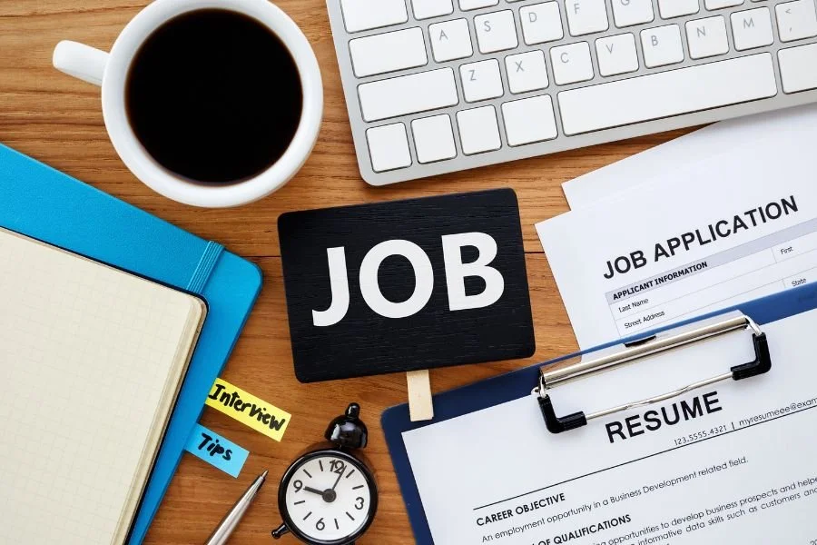 Desk with a cup of coffee, keyboard, job application forms, resume, notebook, sticky notes labeled 'Interview' and 'Tips', pen, small clock, and a 'JOB' sign.