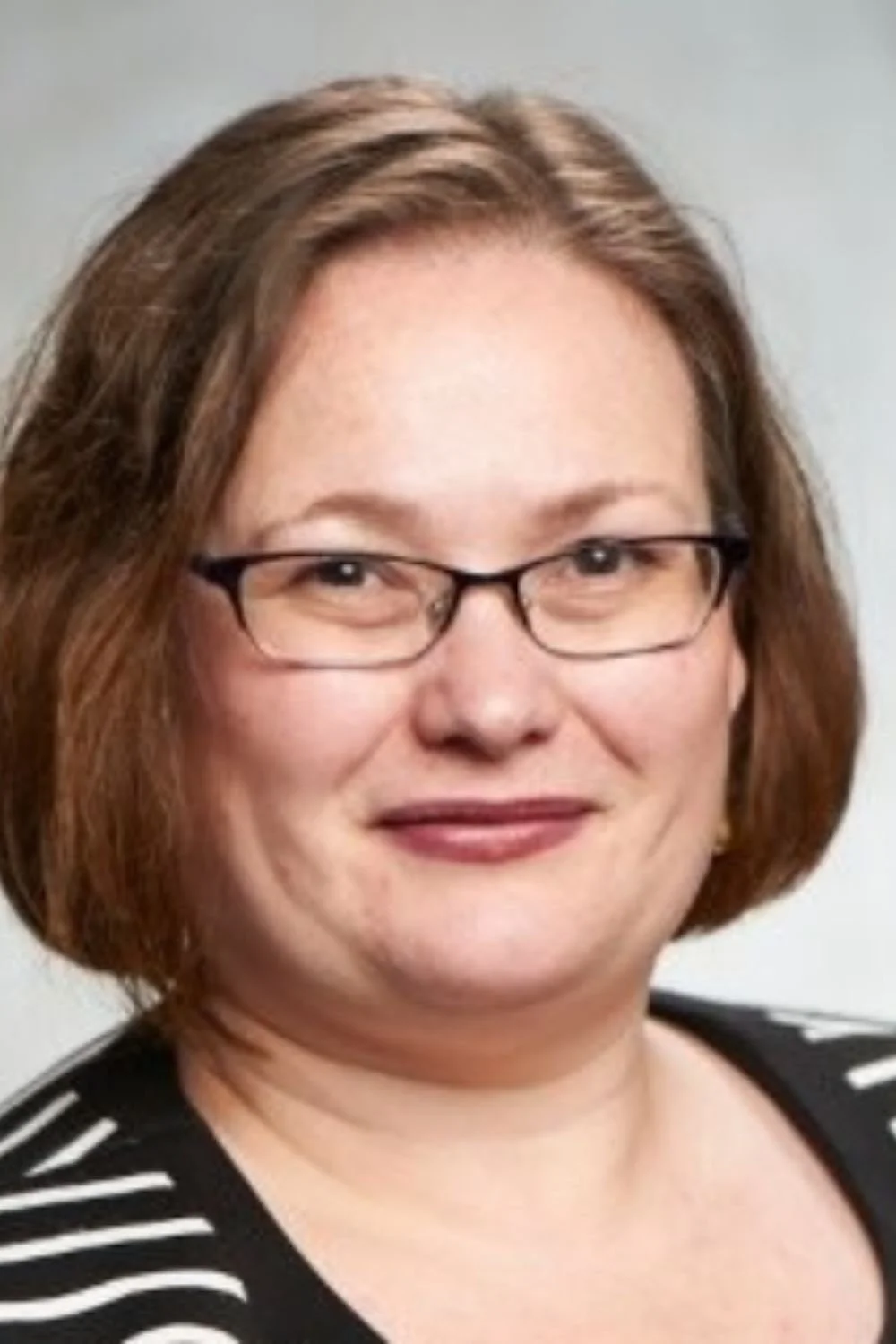 Close-up photo of a woman with glasses, shoulder-length brown hair, wearing a black and white striped top, smiling against a plain background.