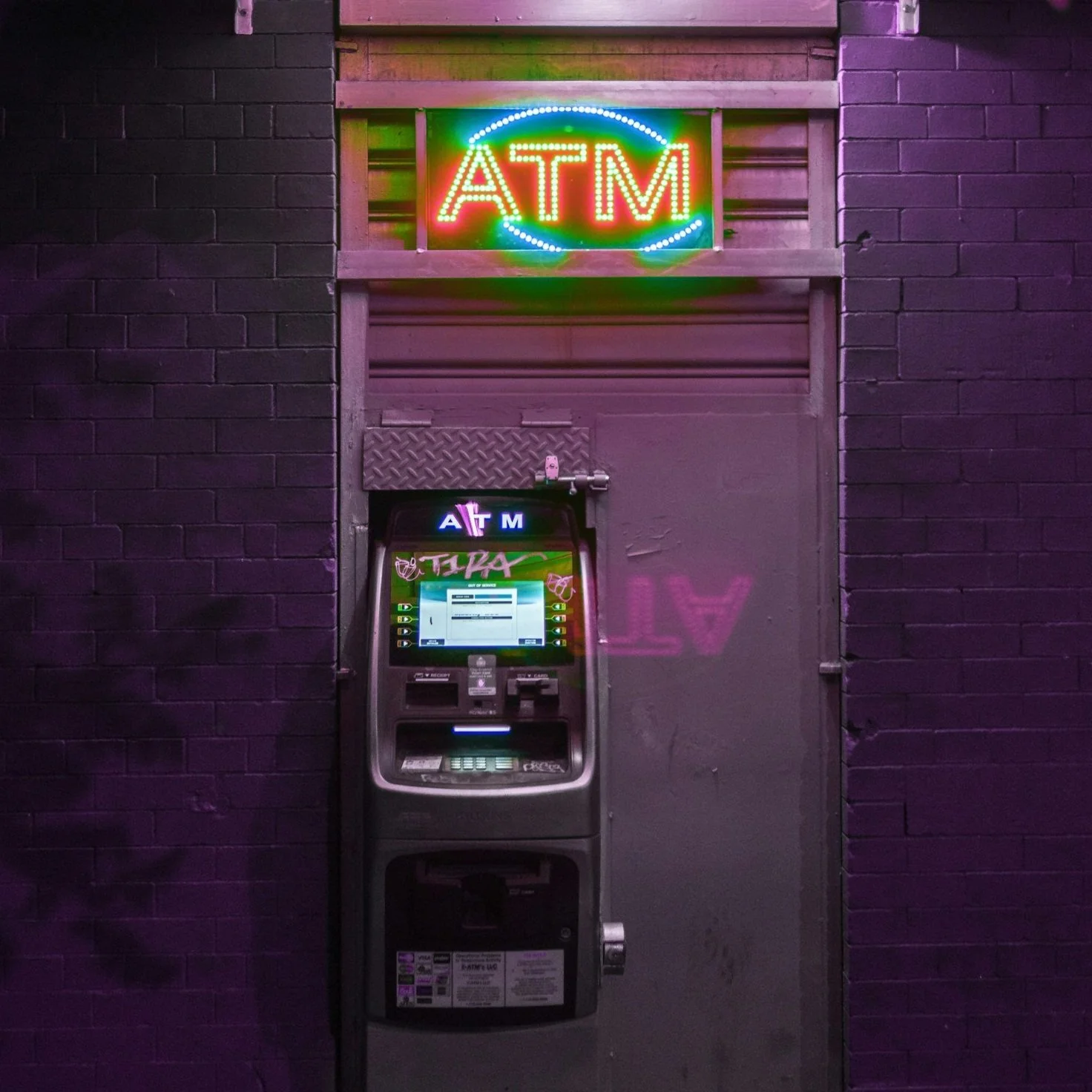 Neon sign displaying ATM above an ATM machine mounted on a dark brick wall, with purple lighting and some graffiti on the wall.