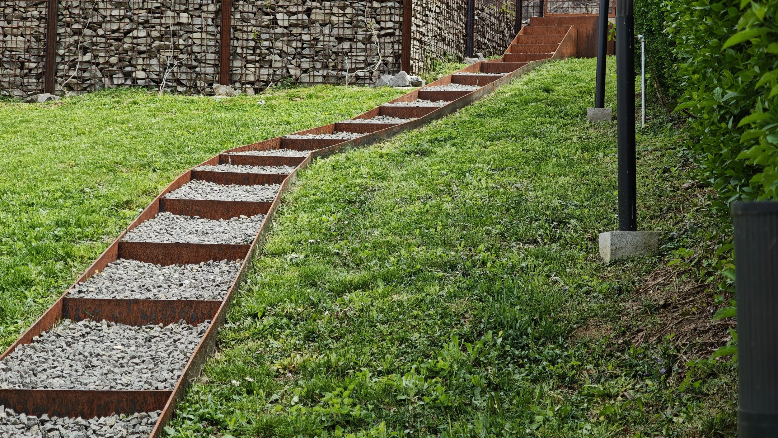 A garden with a rusty metal bed of gravel along a corner, with green grass and plants on either side and a fence in the background.