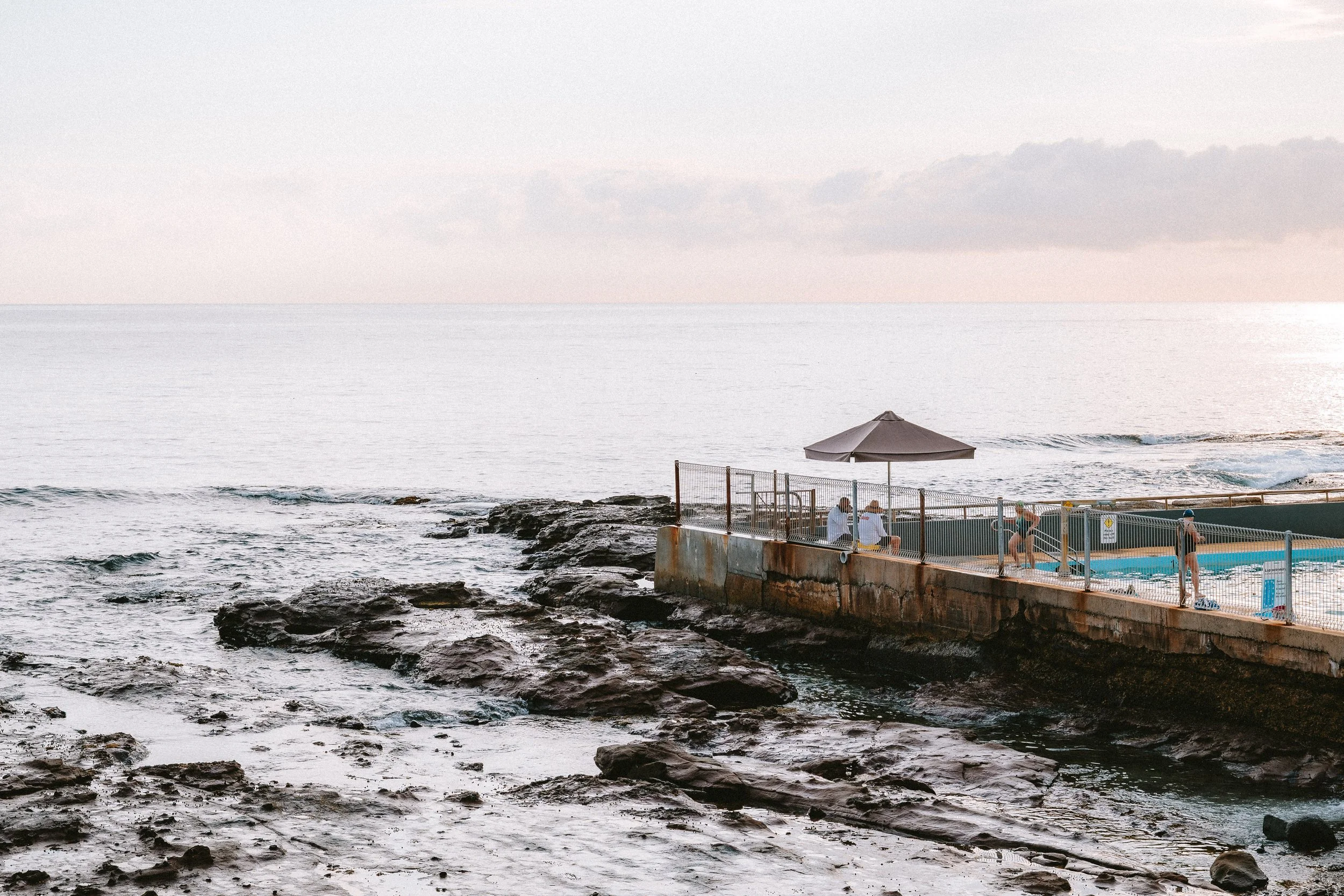 People swimming in an outdoor pool on a rocky coastline at sunset.