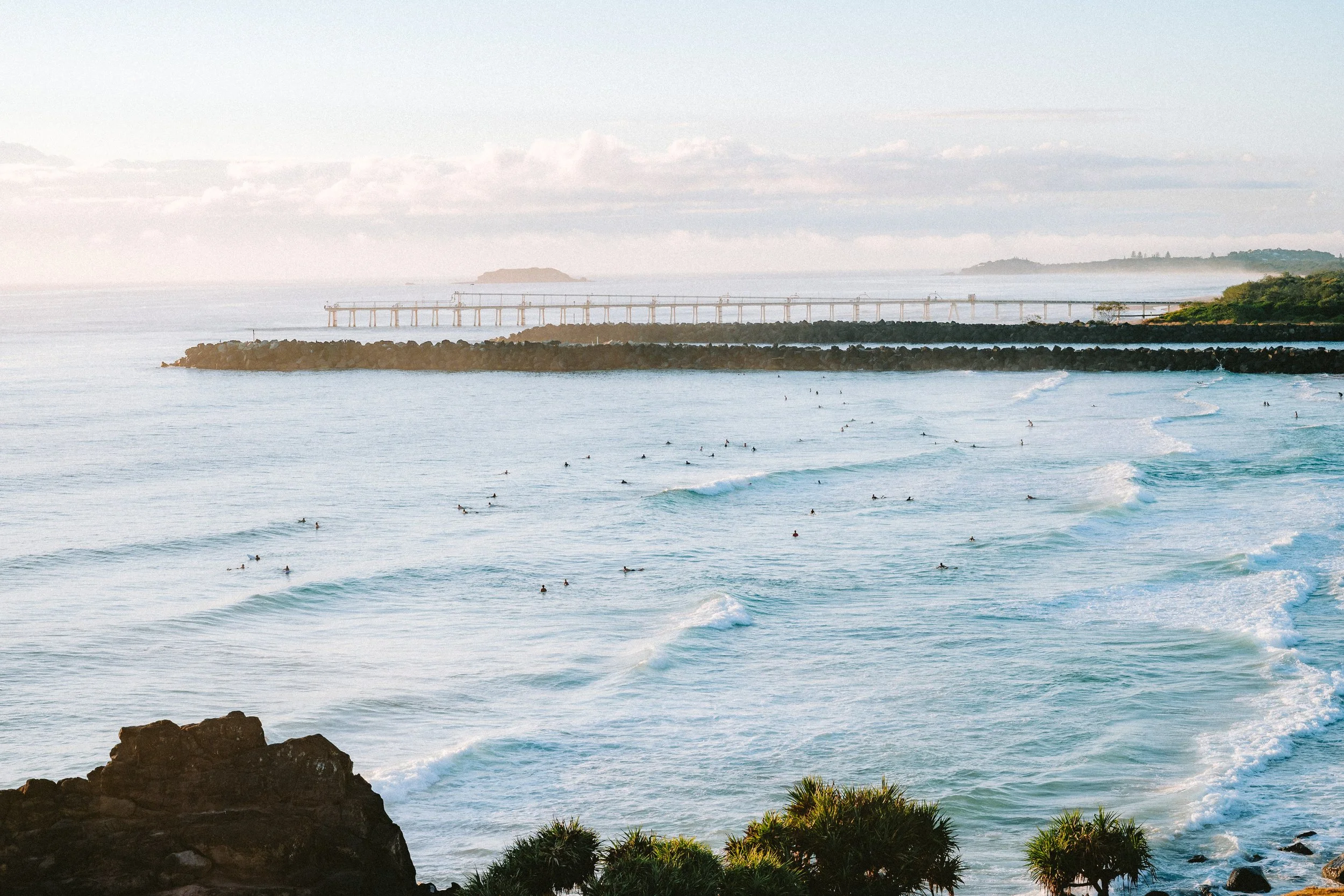 A scenic beach with numerous surfers in the ocean, rocky shoreline in the foreground, a breakwater, and a pier in the distance under a partly cloudy sky.
