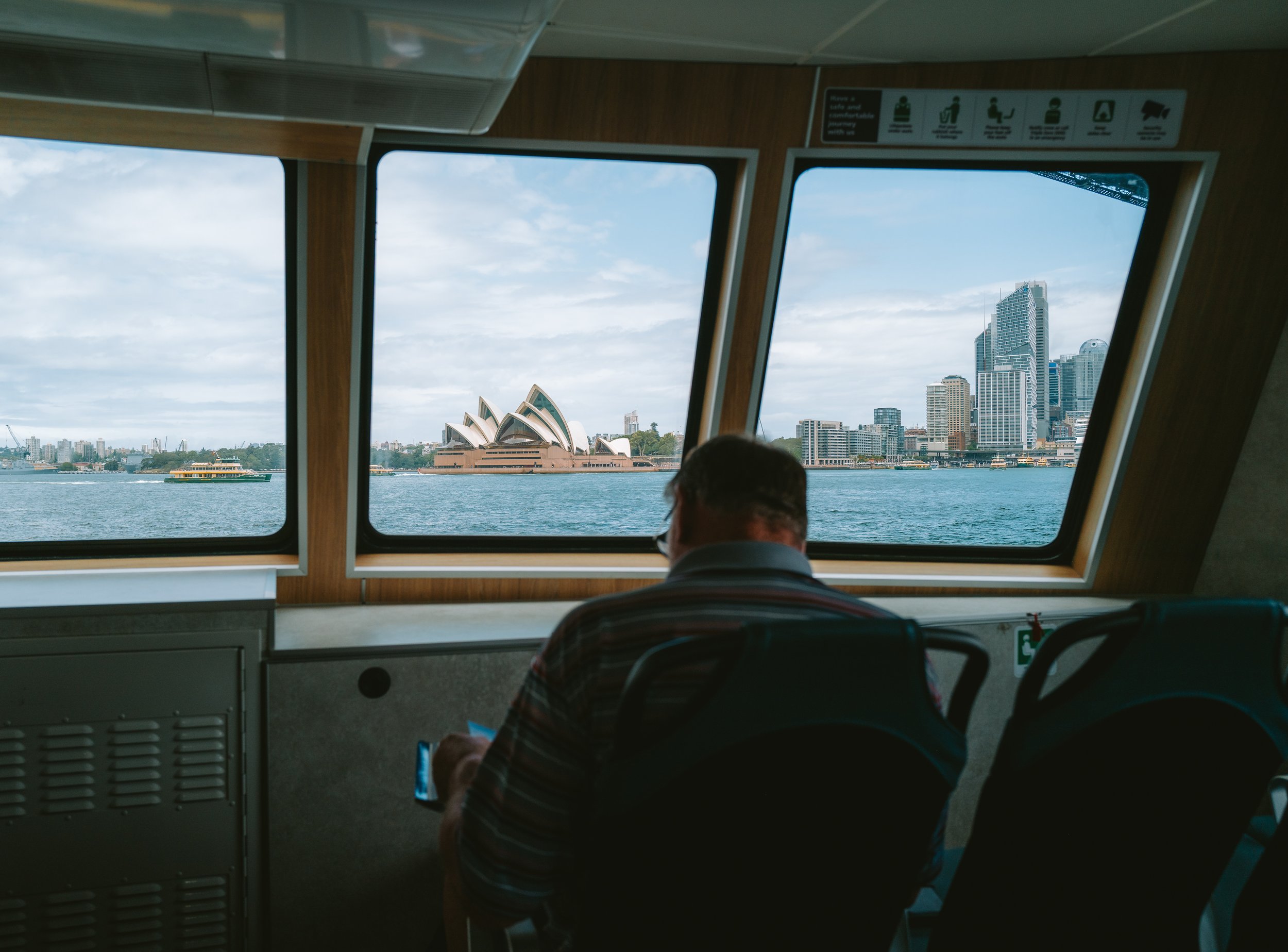 View of Sydney Opera House and city skyline through ferry windows with a person sitting and looking at the view.
