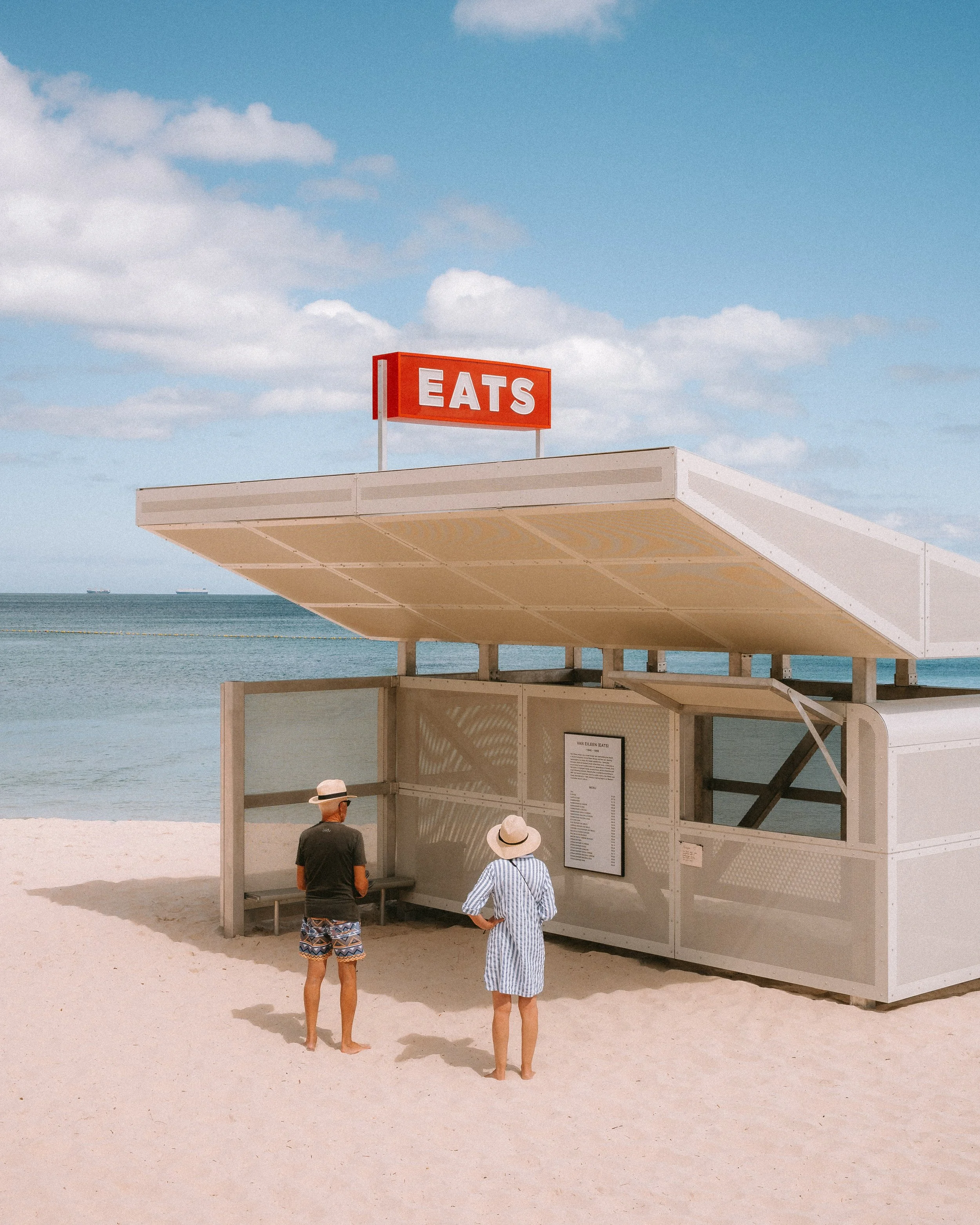 Beachside food kiosk with a red sign that says 'EATS', two people in beachwear and hats standing in front, and a scenic ocean background with a blue sky and scattered clouds.