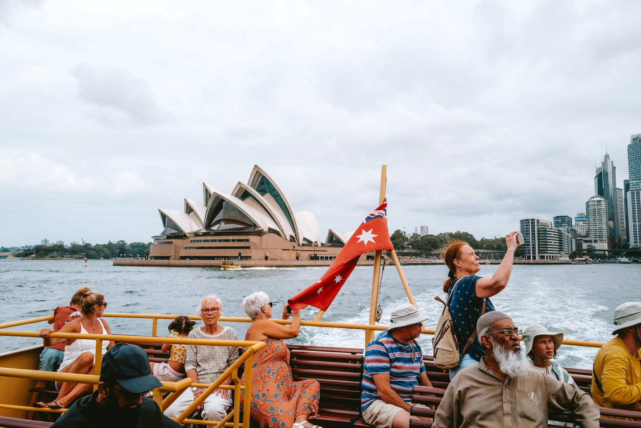 Tourists on a boat ride in Sydney with the Sydney Opera House and city skyline in the background, some taking photos and others enjoying the view.