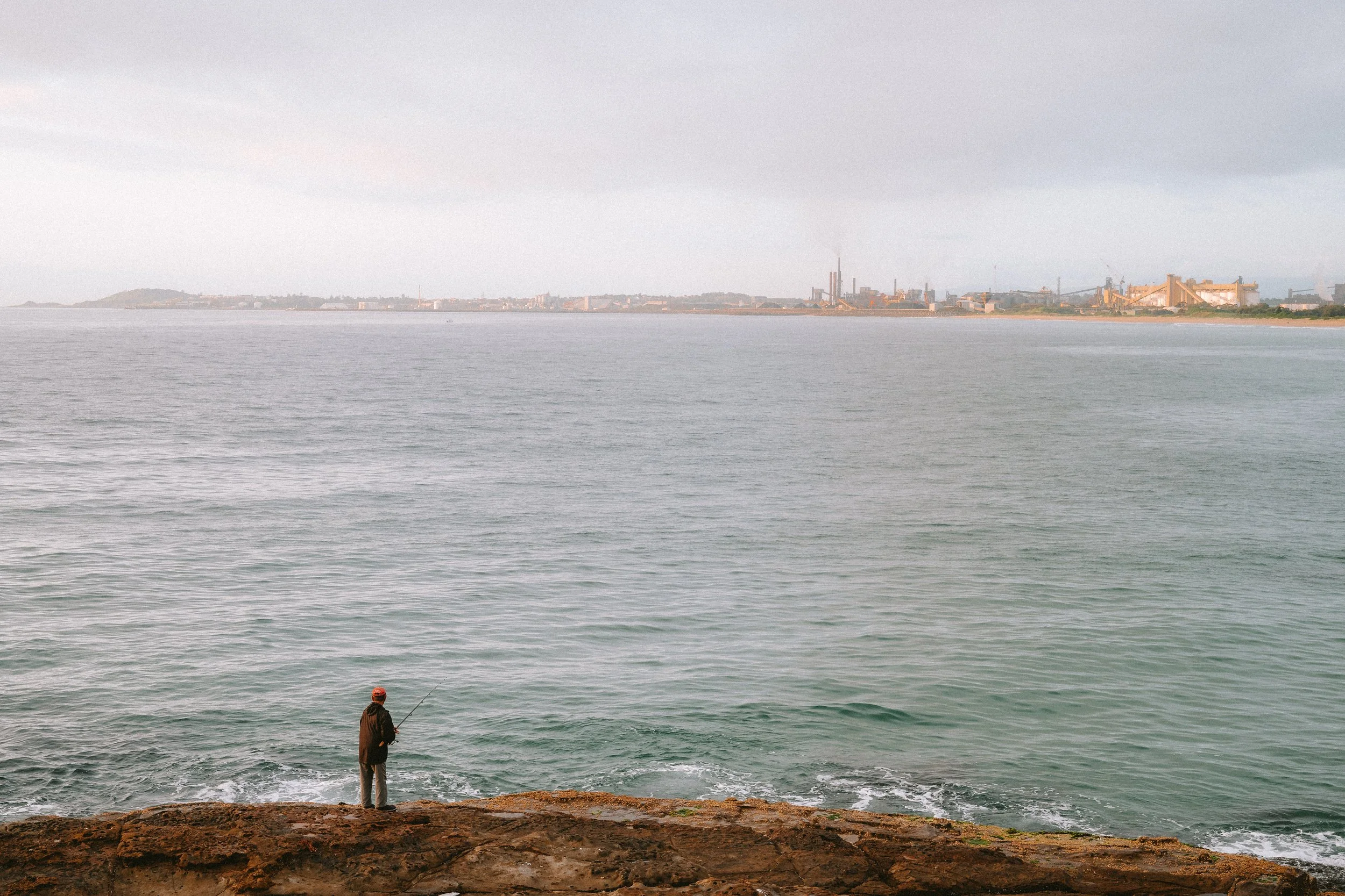 A person fishing on a rocky shoreline with an industrial cityscape across the water in the distance.