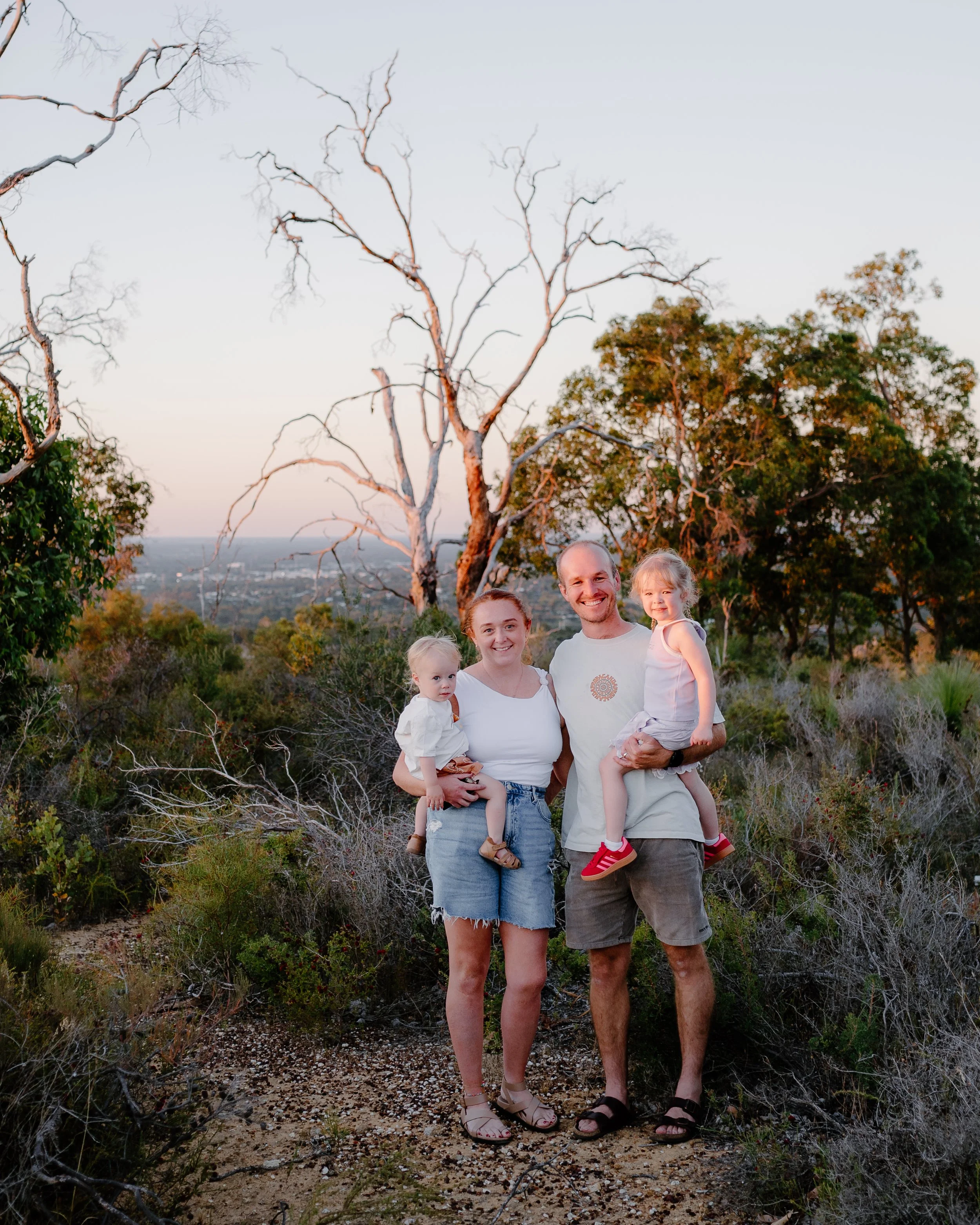 Family of four outdoors during sunset, with trees and a distant city skyline in the background.