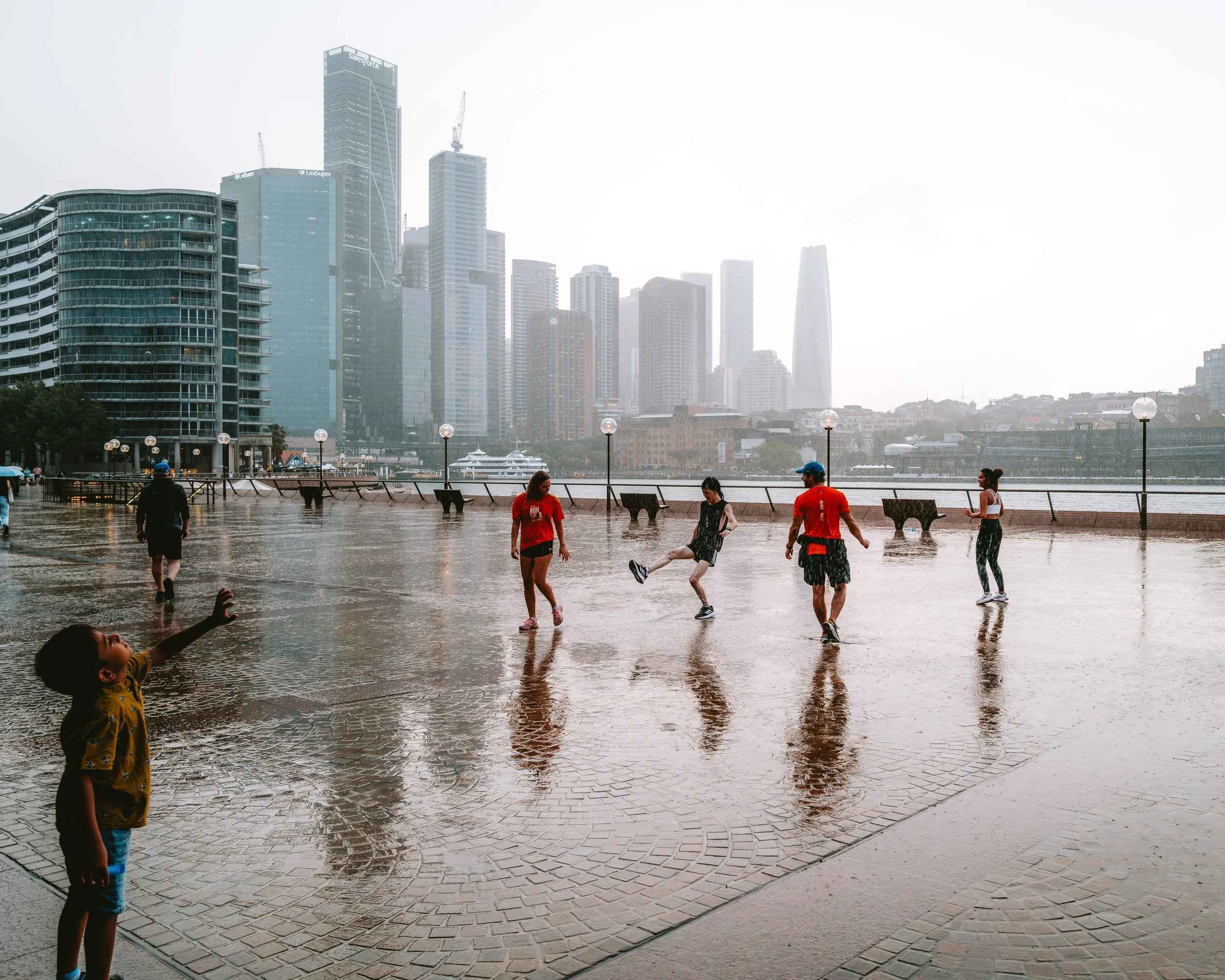 Children and adults playing on a wet promenade with city skyscrapers in the background during a rainy day.