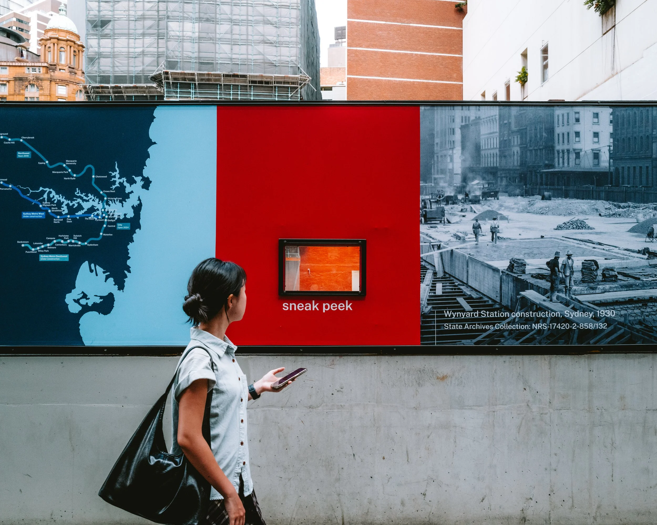A woman walking past a large informational display about the construction of Wynyard Station in Sydney in 1930. The display features a map of the Sydney area, a black and white photograph of the construction site, and a small window labeled 'sneak pe