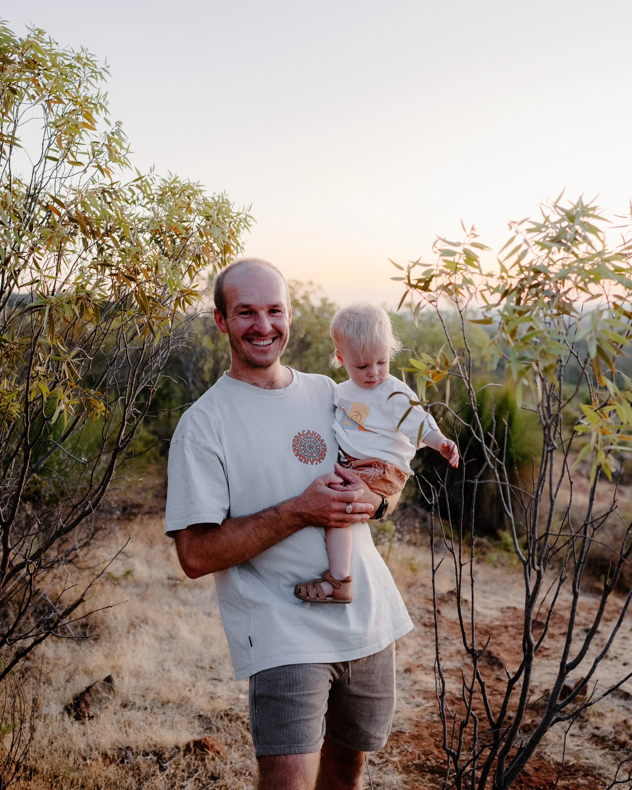 A man smiling while holding a young boy outdoors during sunset, surrounded by shrubs and dry grass.