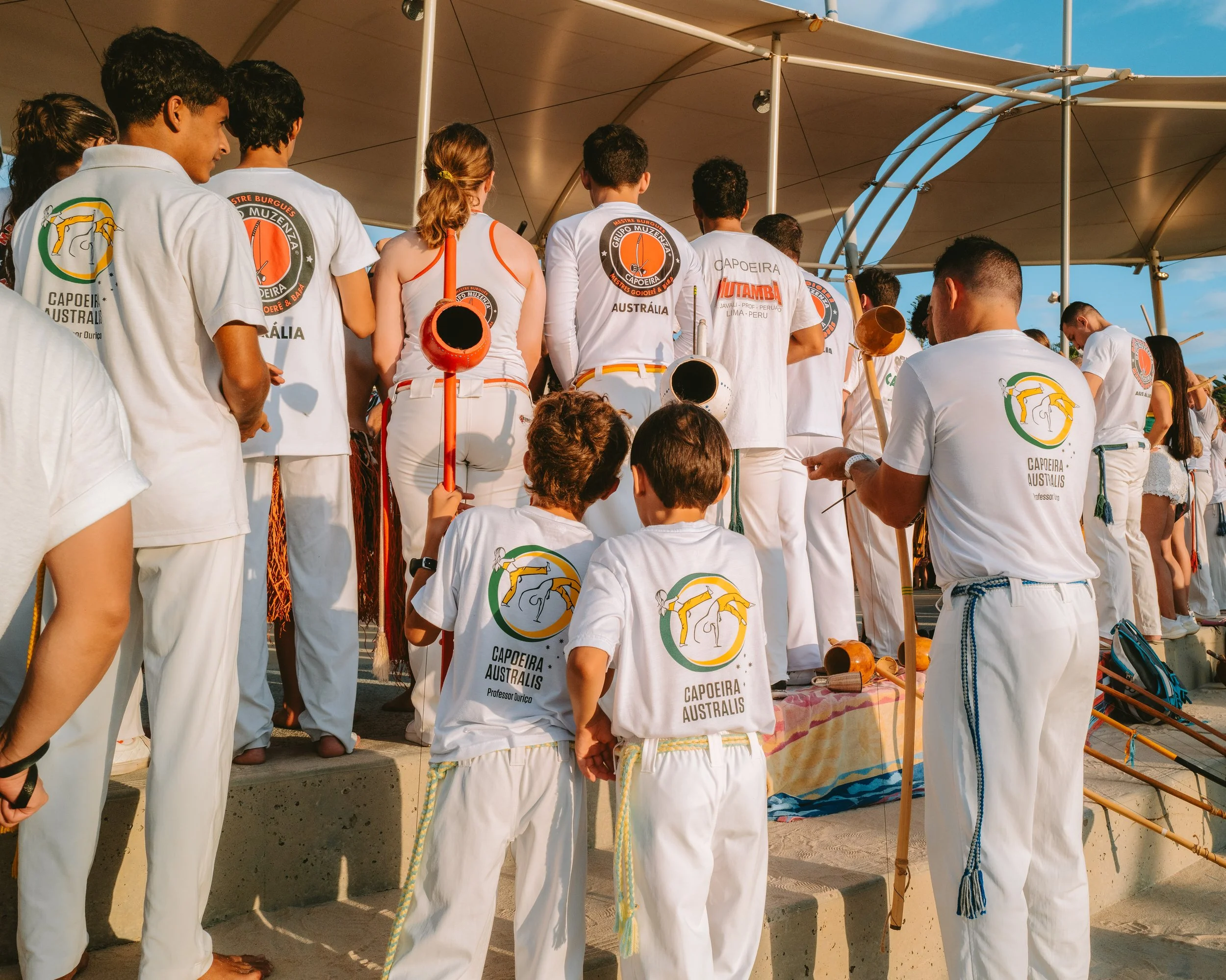 Group of people dressed in white clothing, standing on steps under a canopy, participating in a capoeira event with some holding traditional instruments.