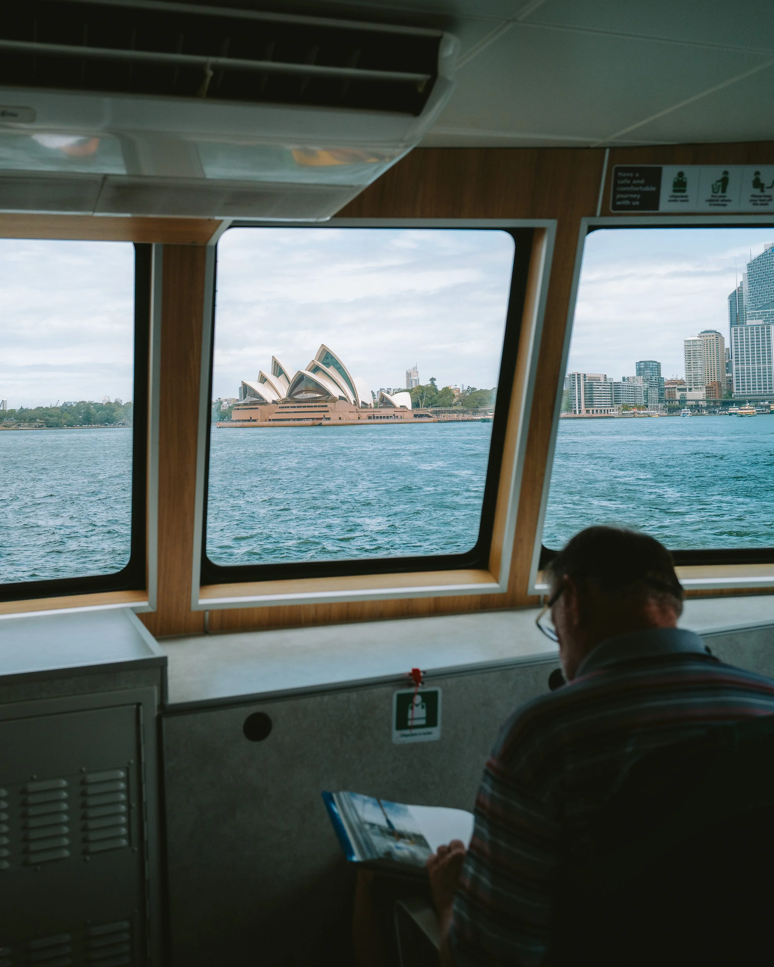 View of the Sydney Opera House and Sydney skyline through ferry window, with a person reading a brochure in the foreground.
