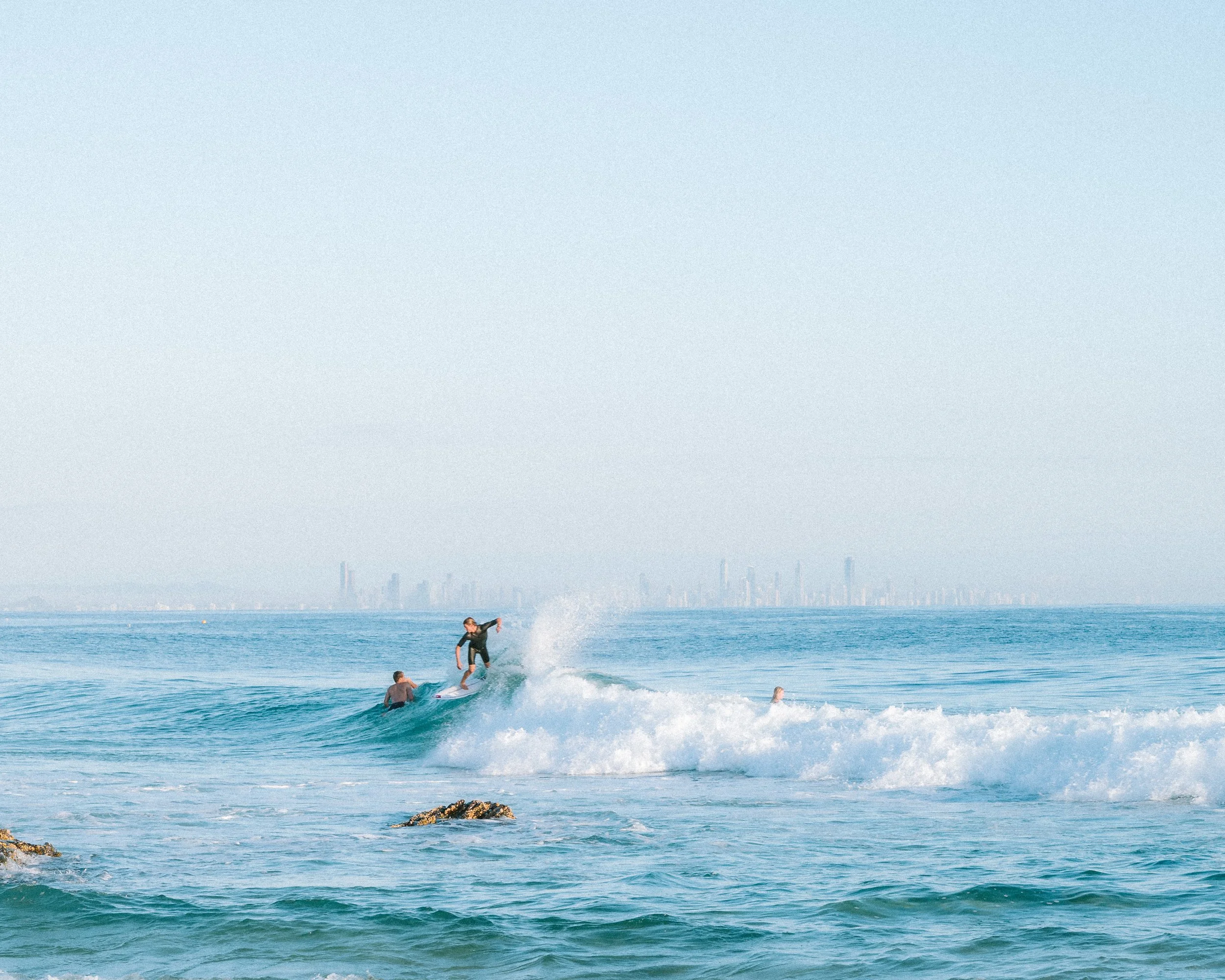 A surfer riding a wave in the ocean with a city skyline in the background.