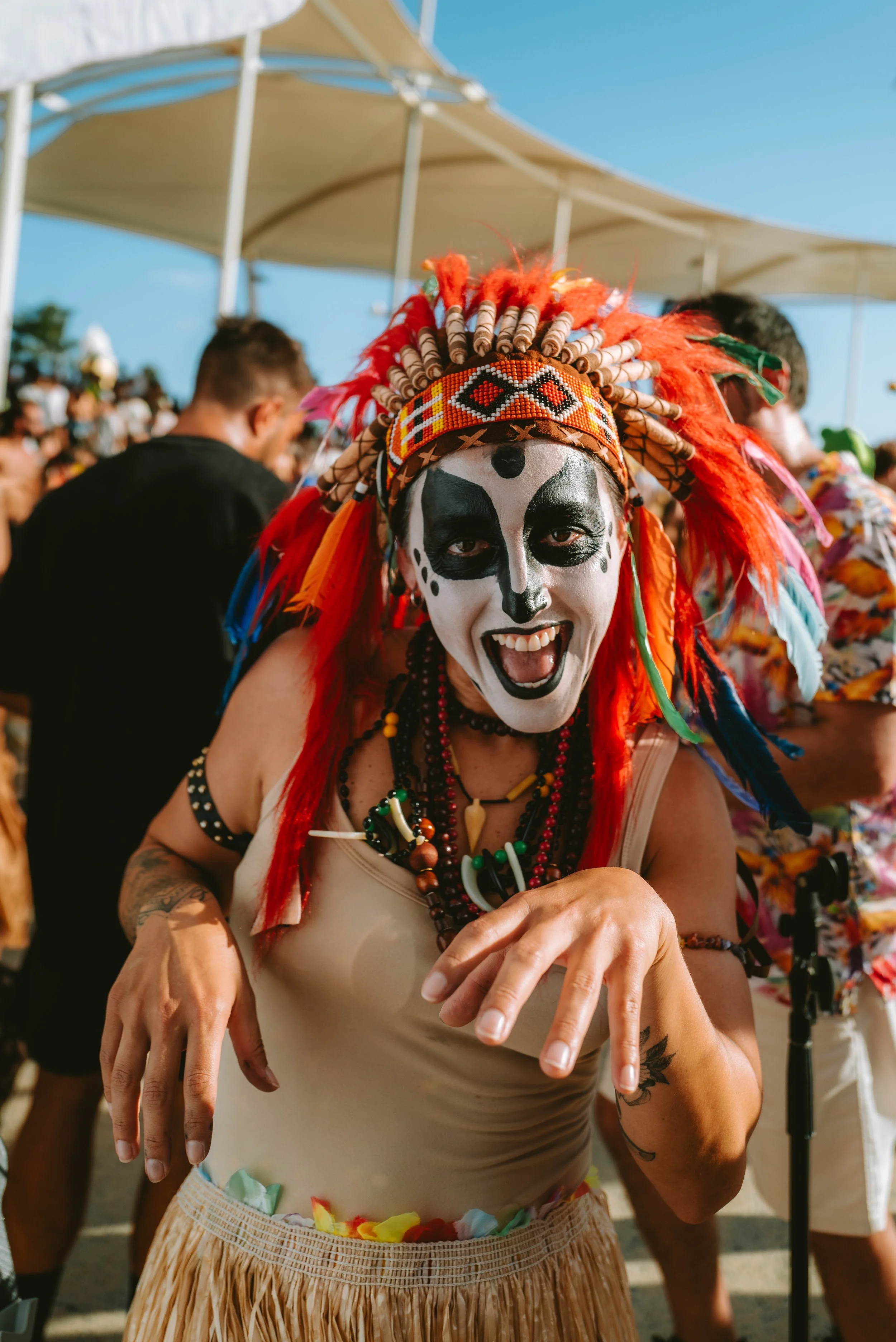 Person dressed in colorful tribal costume with face paint, feathers, and beads, smiling at a festival or celebration.