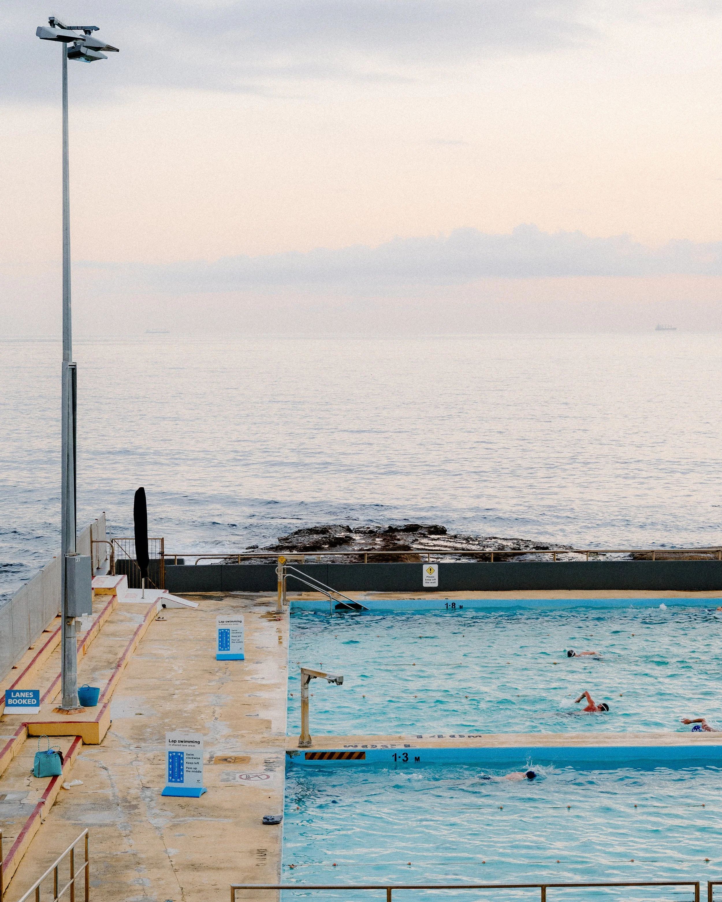 An outdoor swimming pool near the ocean with swimmers in the water, pool depth markers, and a sign indicating lanes are booked, with a rocky shoreline and ships visible in the distance.