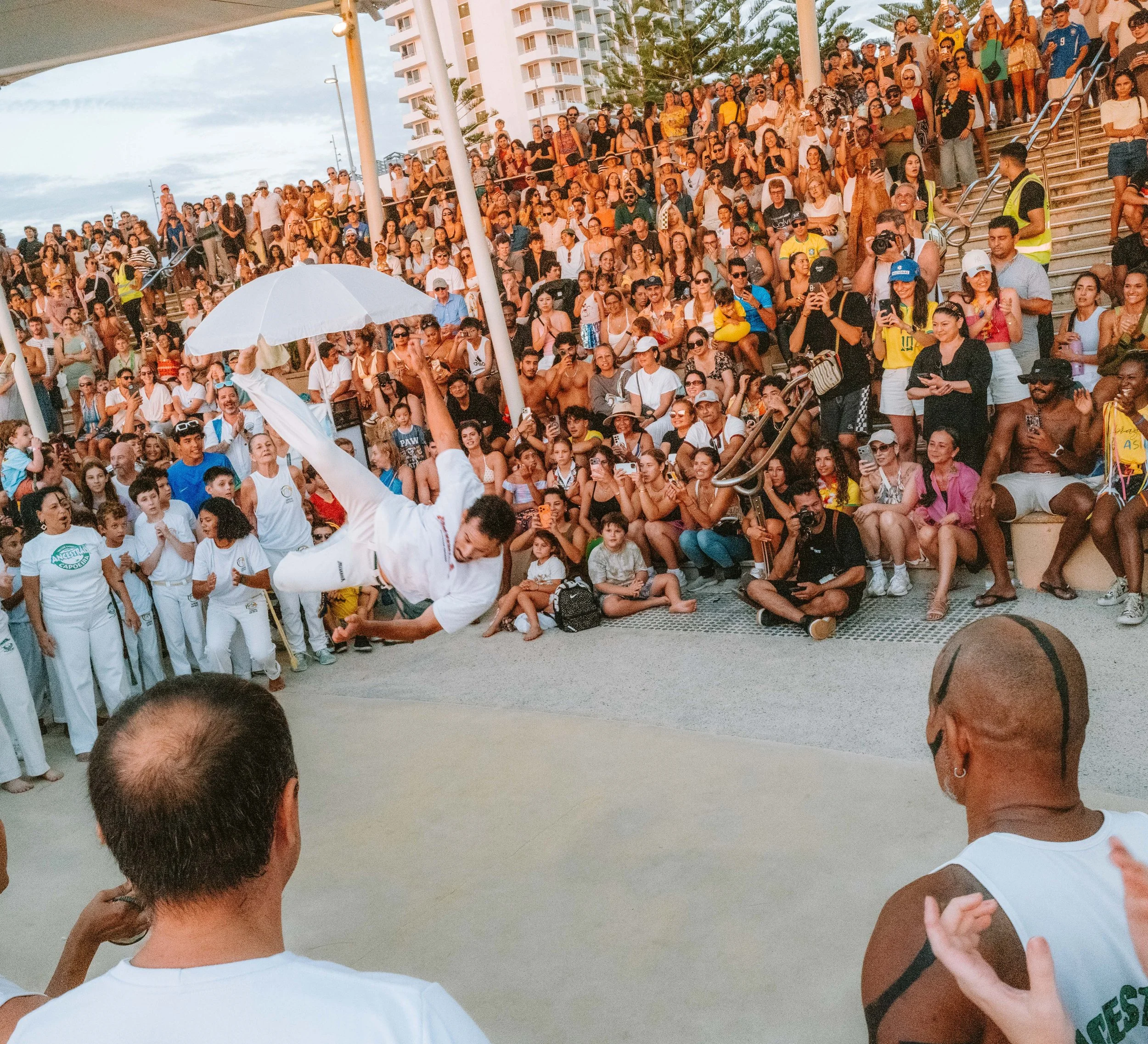 Performer in white clothing mid-air doing a breakdancing move with an umbrella in front of a large crowd at an outdoor event.