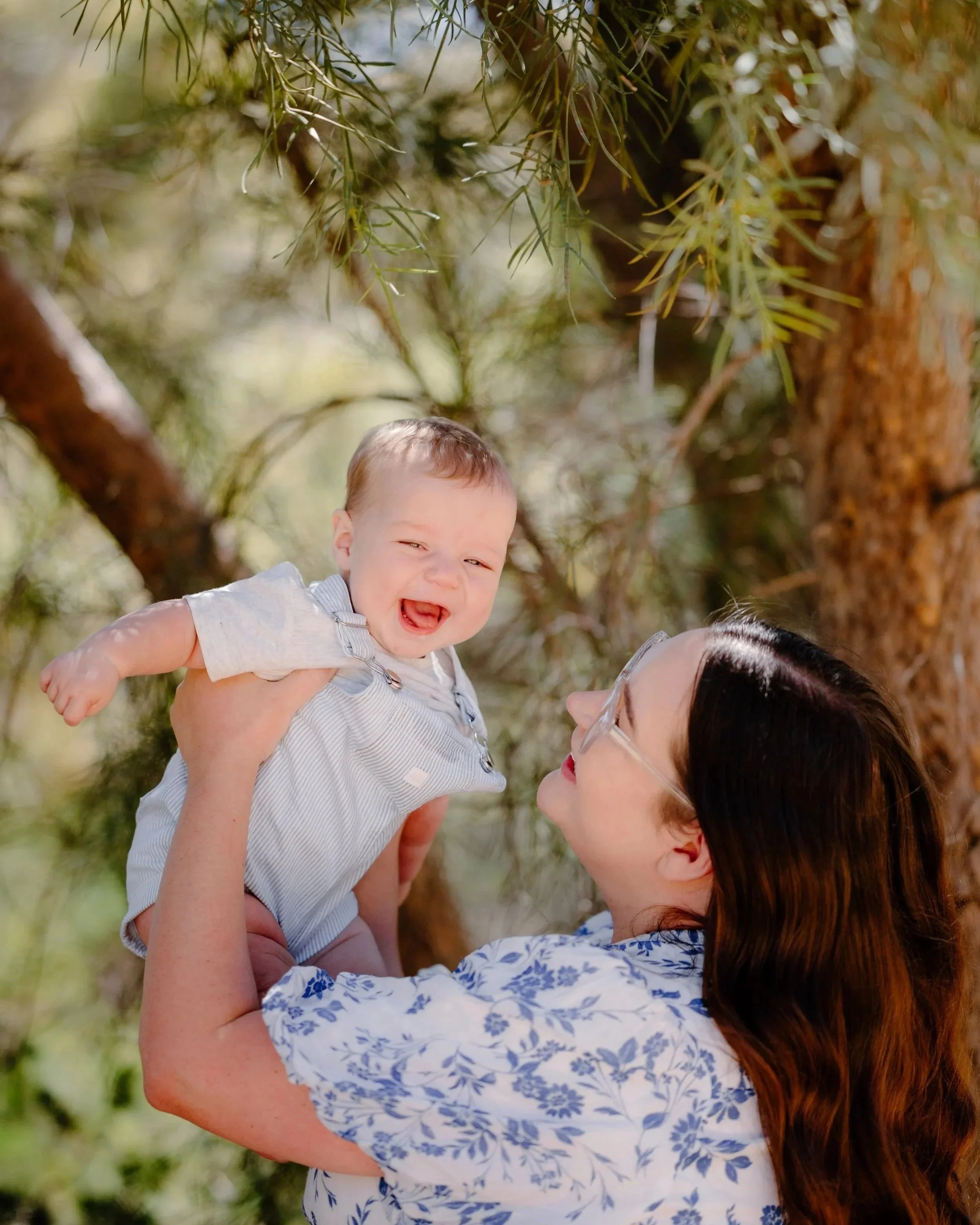 A woman with red hair and sunglasses holding a laughing baby boy in a branch of a tree with green foliage.