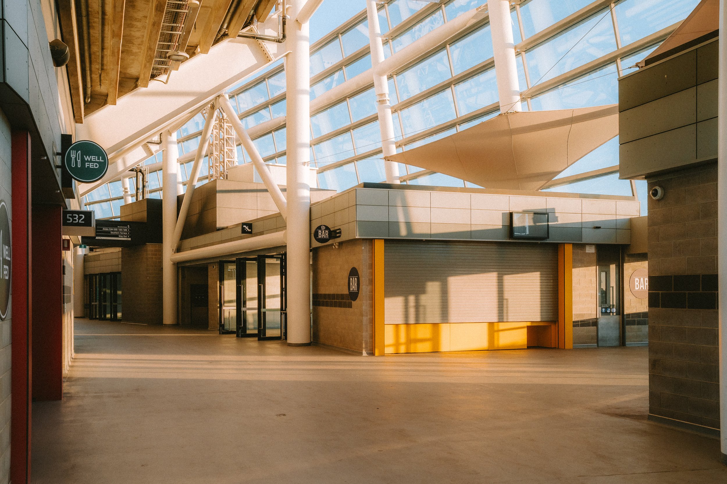 Empty indoor commercial space with closed storefronts, glass doors, and sunlight casting shadows on the floor, with some signs indicating a bar and food service.