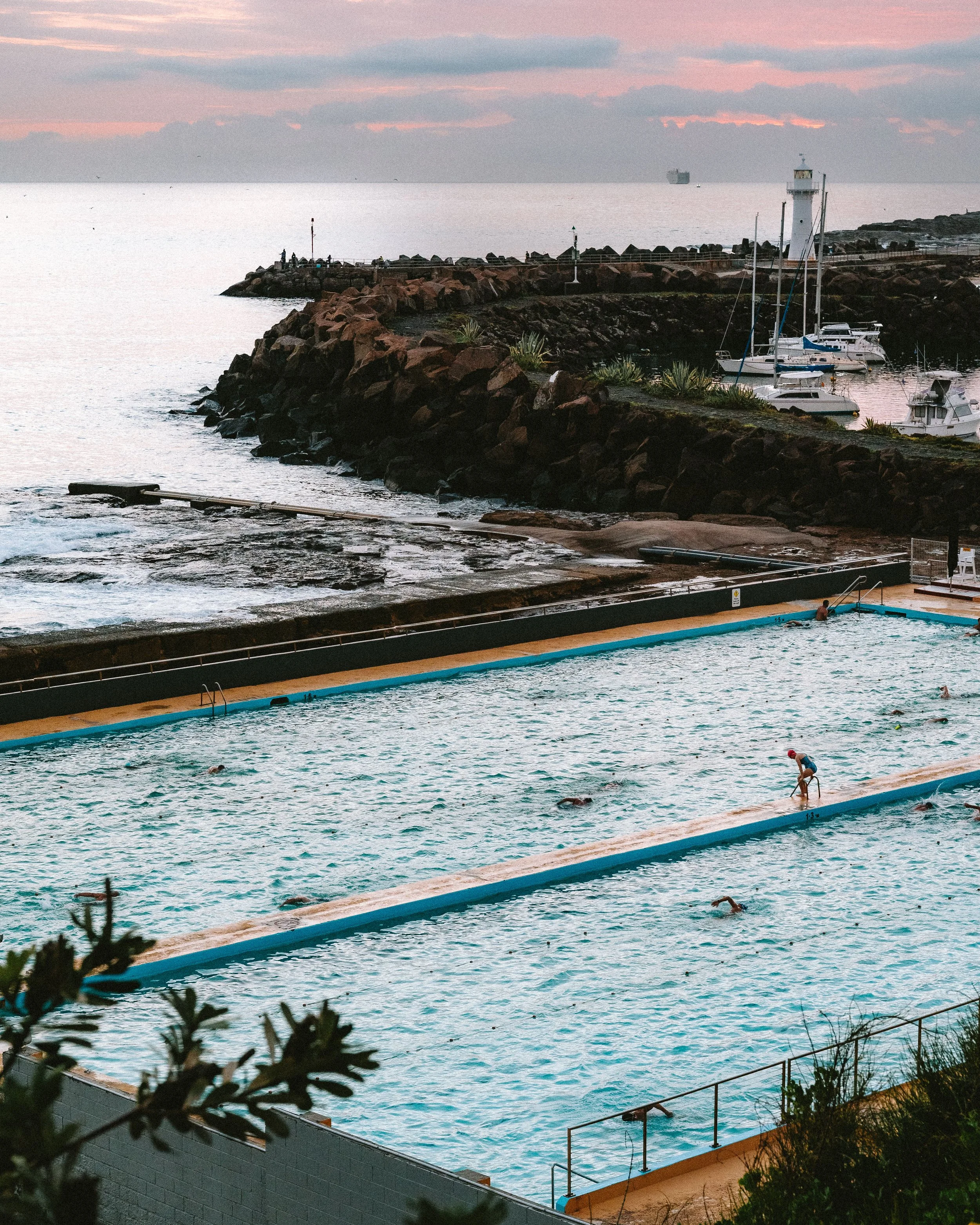 Outdoor swimming pool with several swimmers, surrounded by a fence and plants, with a rocky shoreline, sailboats docked nearby, and a lighthouse in the distance at sunset.