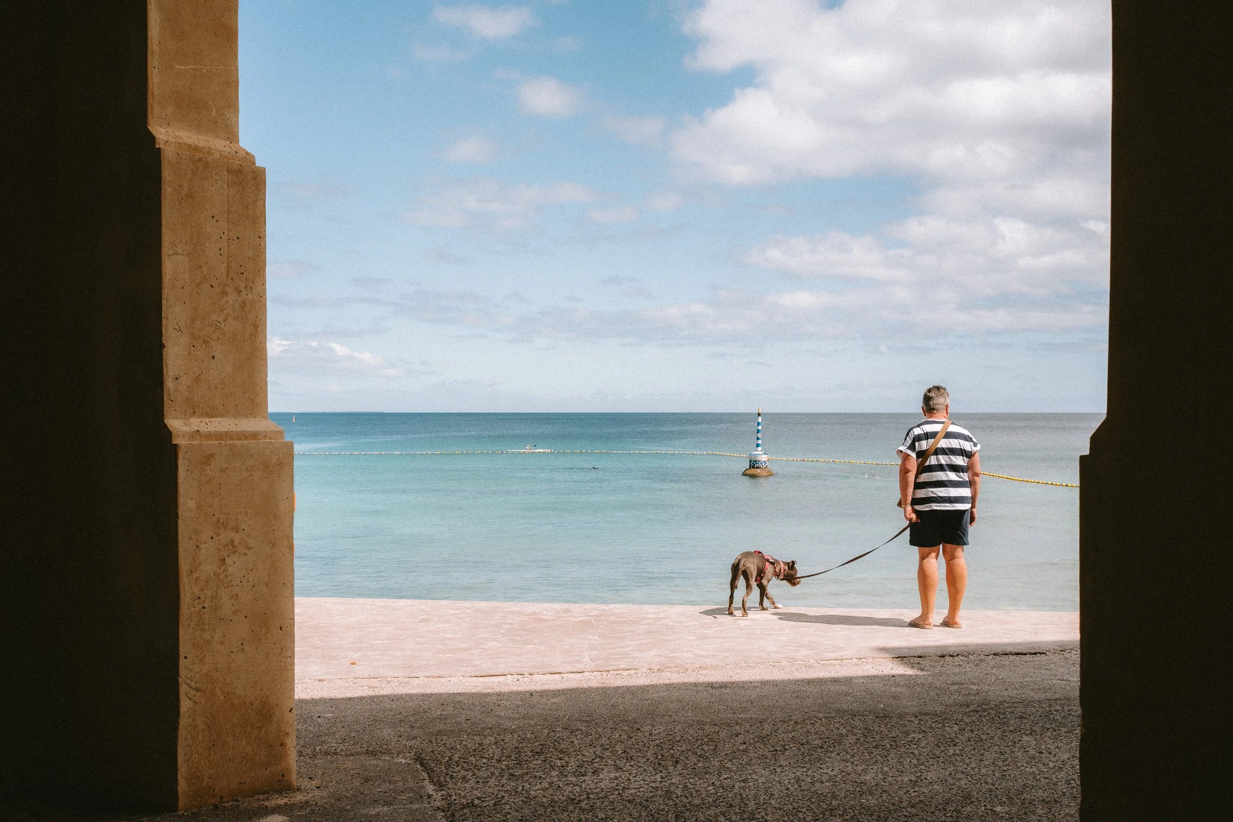 A woman walking her dog on a beach viewed from inside a structure or tunnel, with the sea and sky in the background.