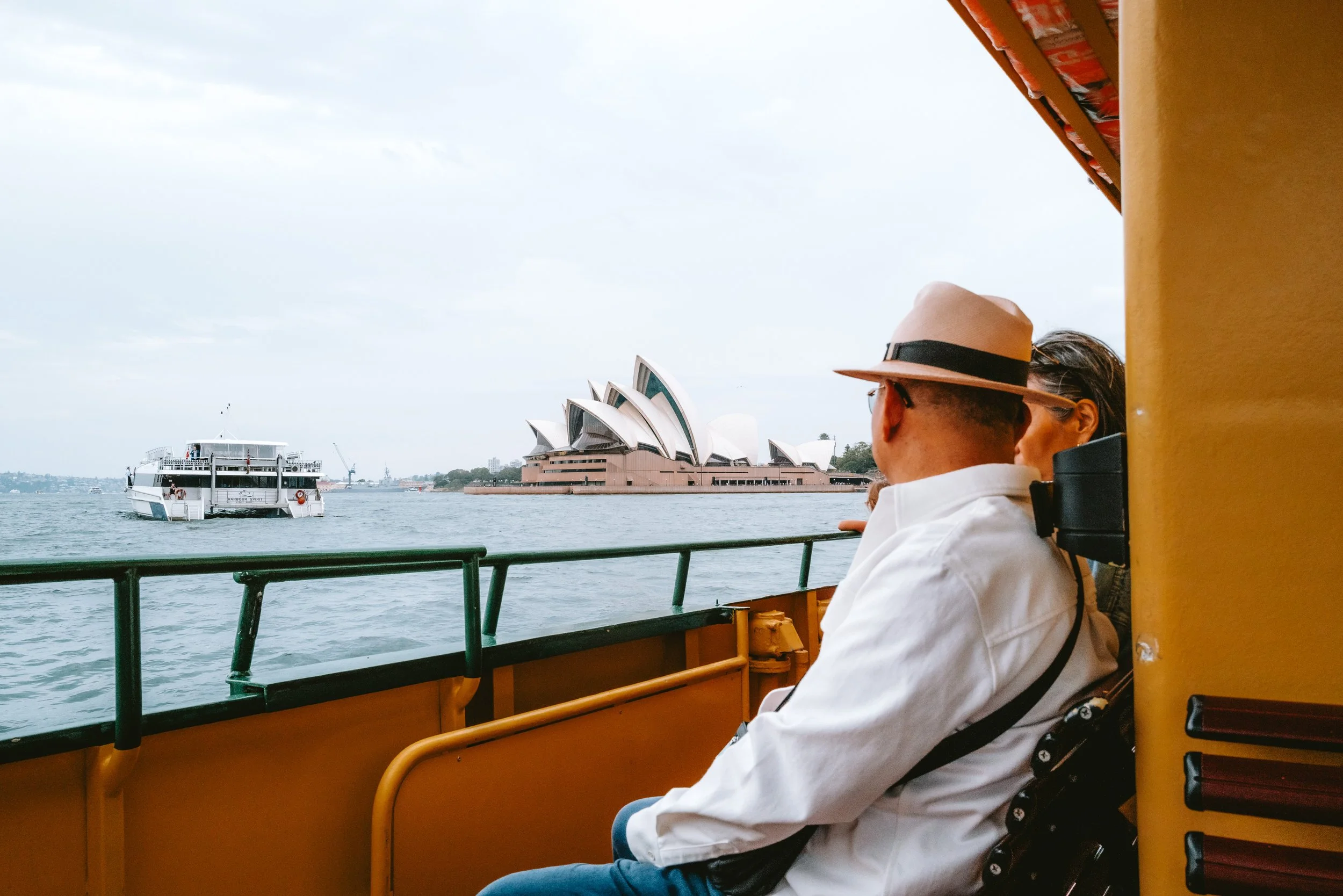 People sitting on a ferry boat enjoying a view of the Sydney Opera House in Australia with water and other boats.