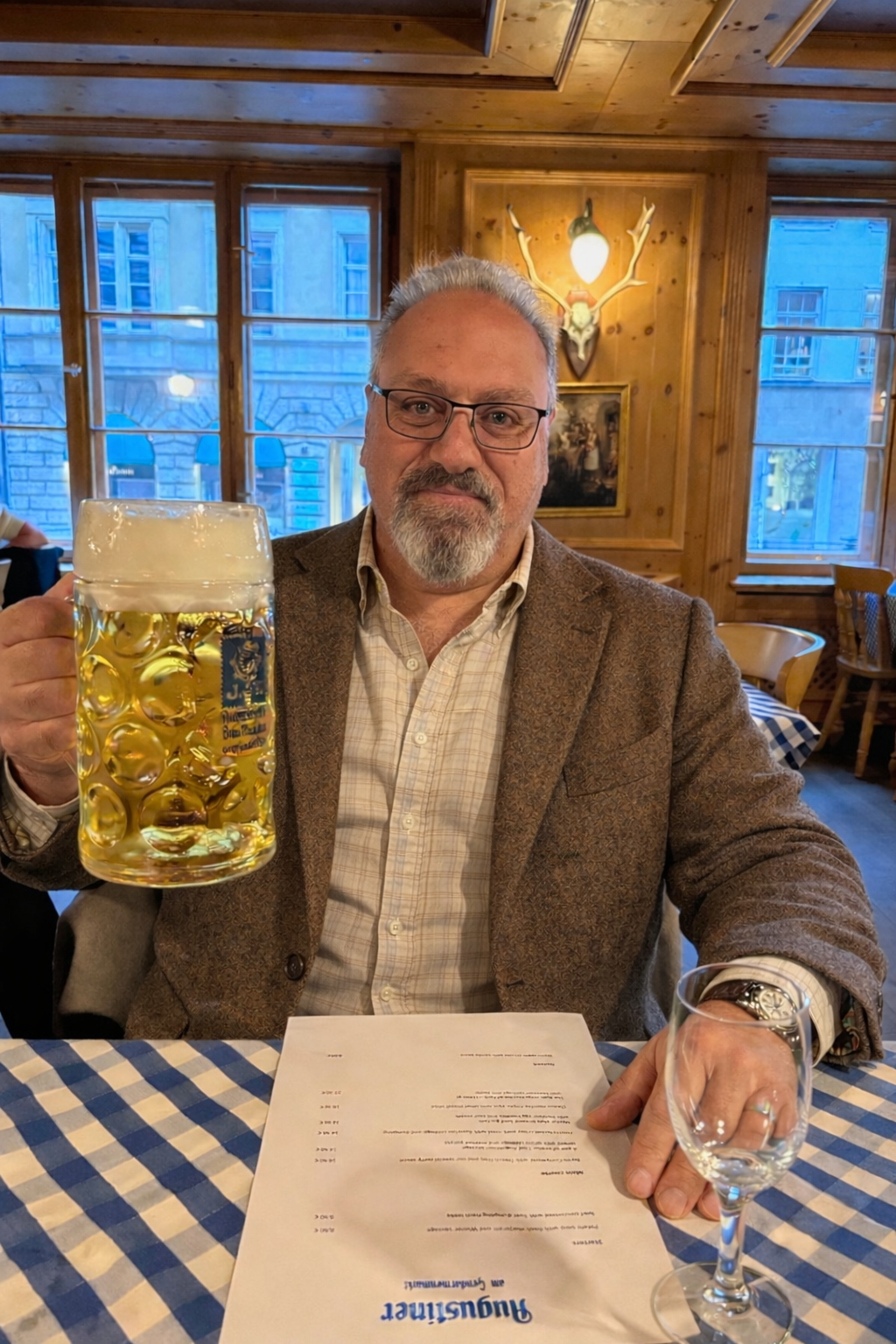 A man with glasses and a beard sitting at a table in a wooden restaurant, holding a large beer mug. There is a menu on the table, and he is looking at the camera with a slight smile.