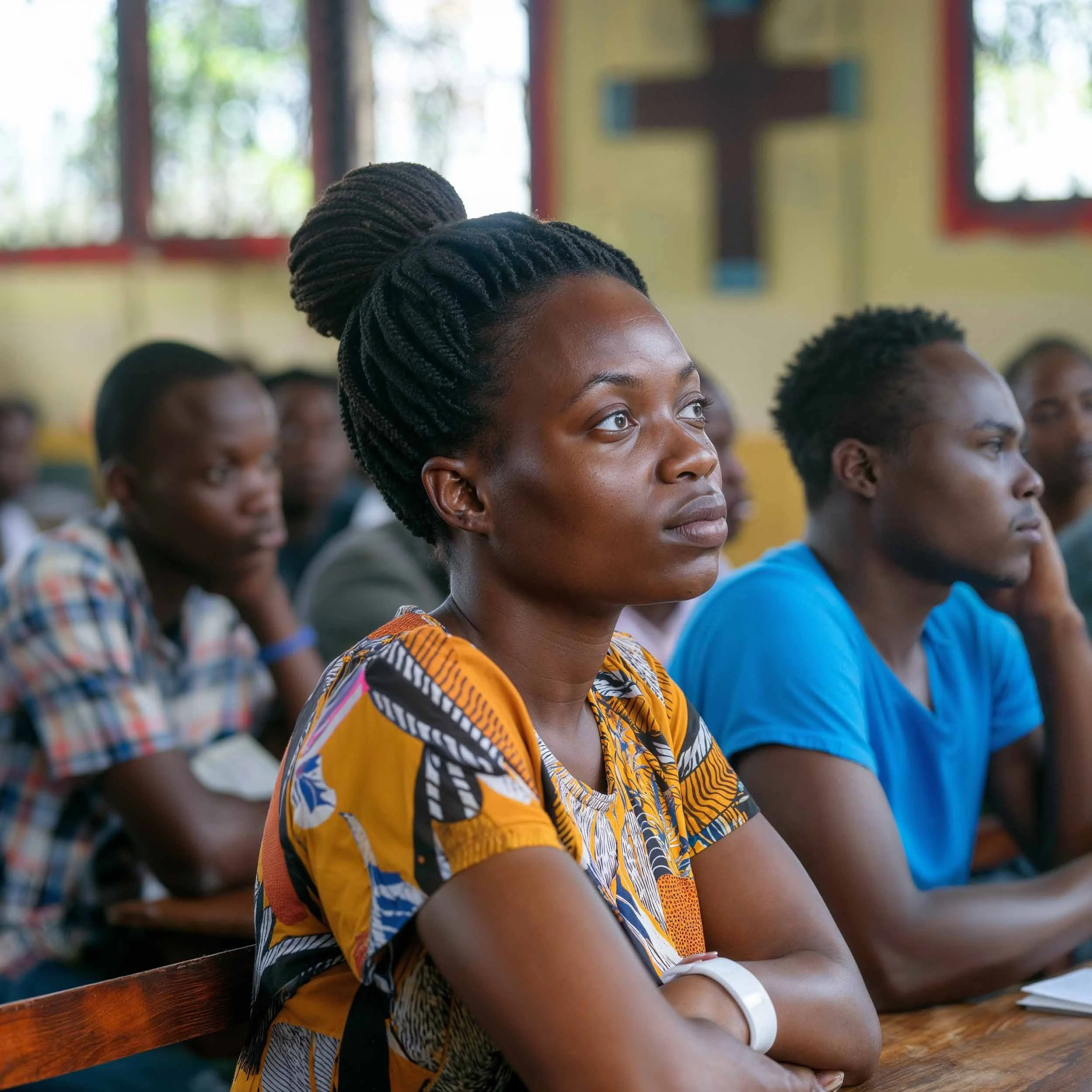 A group of people attentively listening in a classroom, with a woman in a colorful top in the foreground and a cross on the wall in the background.