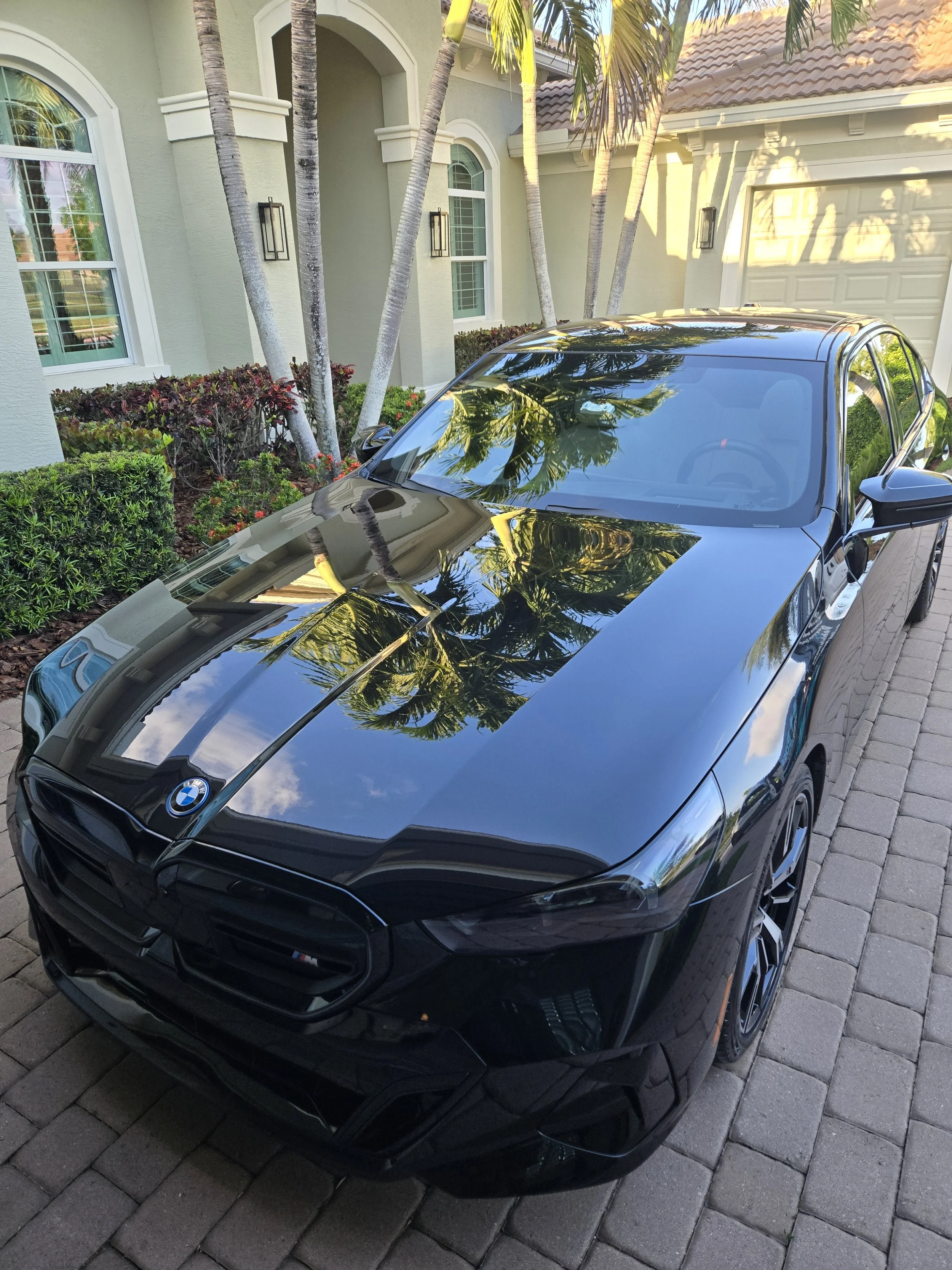 A black BMW car parked on a brick driveway in front of a house with a light green exterior and palm trees.