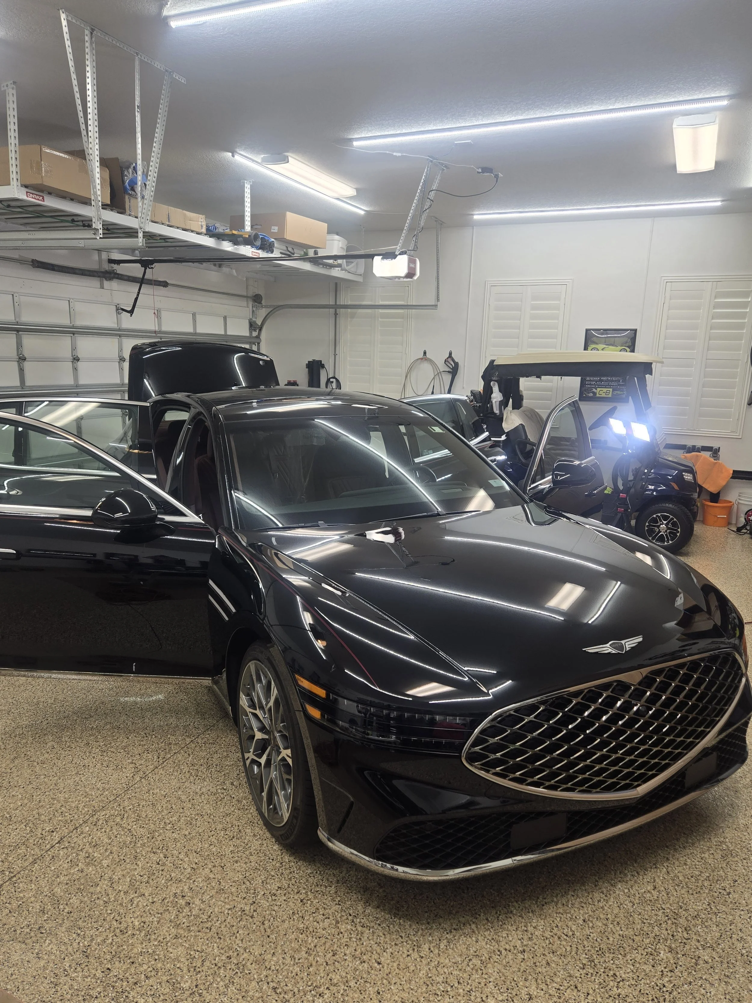 A garage with a black luxury car, a golf cart, and various vehicles, with a garage door, ceiling lights, shelved storage, and a speckled floor.