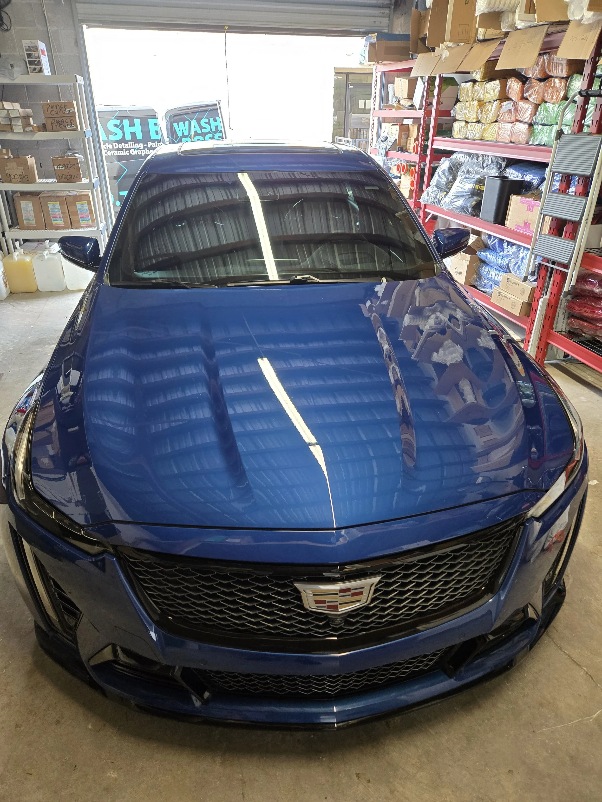Front view of a blue Cadillac car parked inside a garage or warehouse, with shelves on the sides stock with various items and boxes, and bright light coming from open garage doors behind the car.