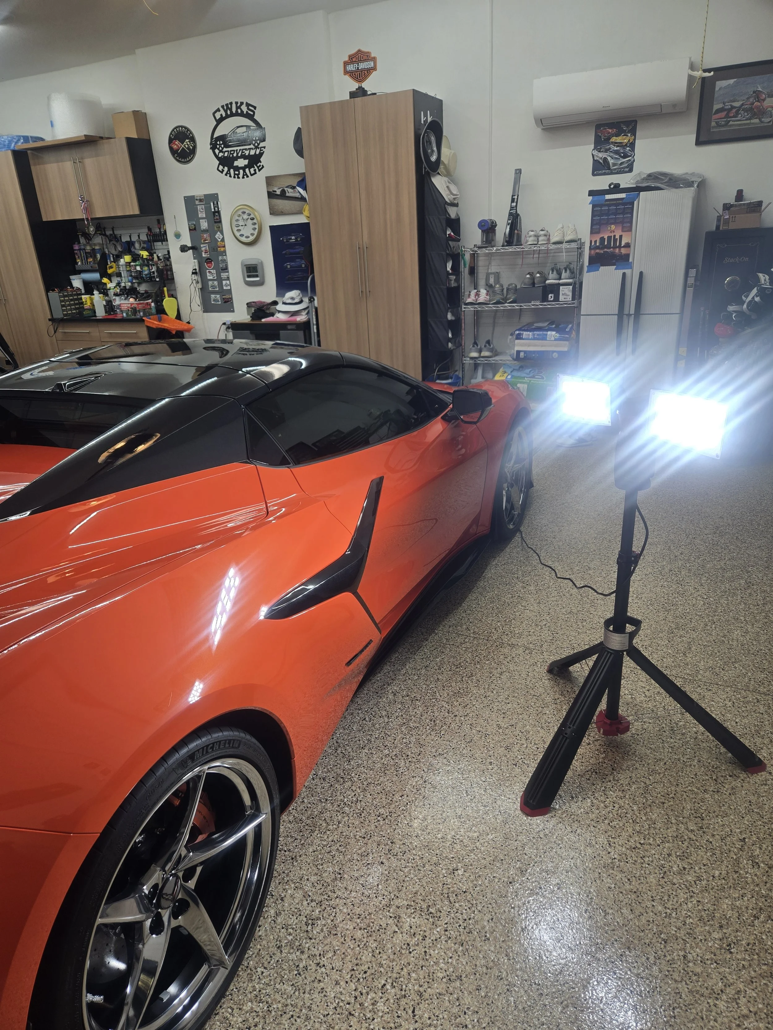 In a garage, there is a bright orange sports car with black accents and large rims in the foreground. The background shows shelves and cabinets, car-related posters, and a light stand with bright LEDs lighting the scene.