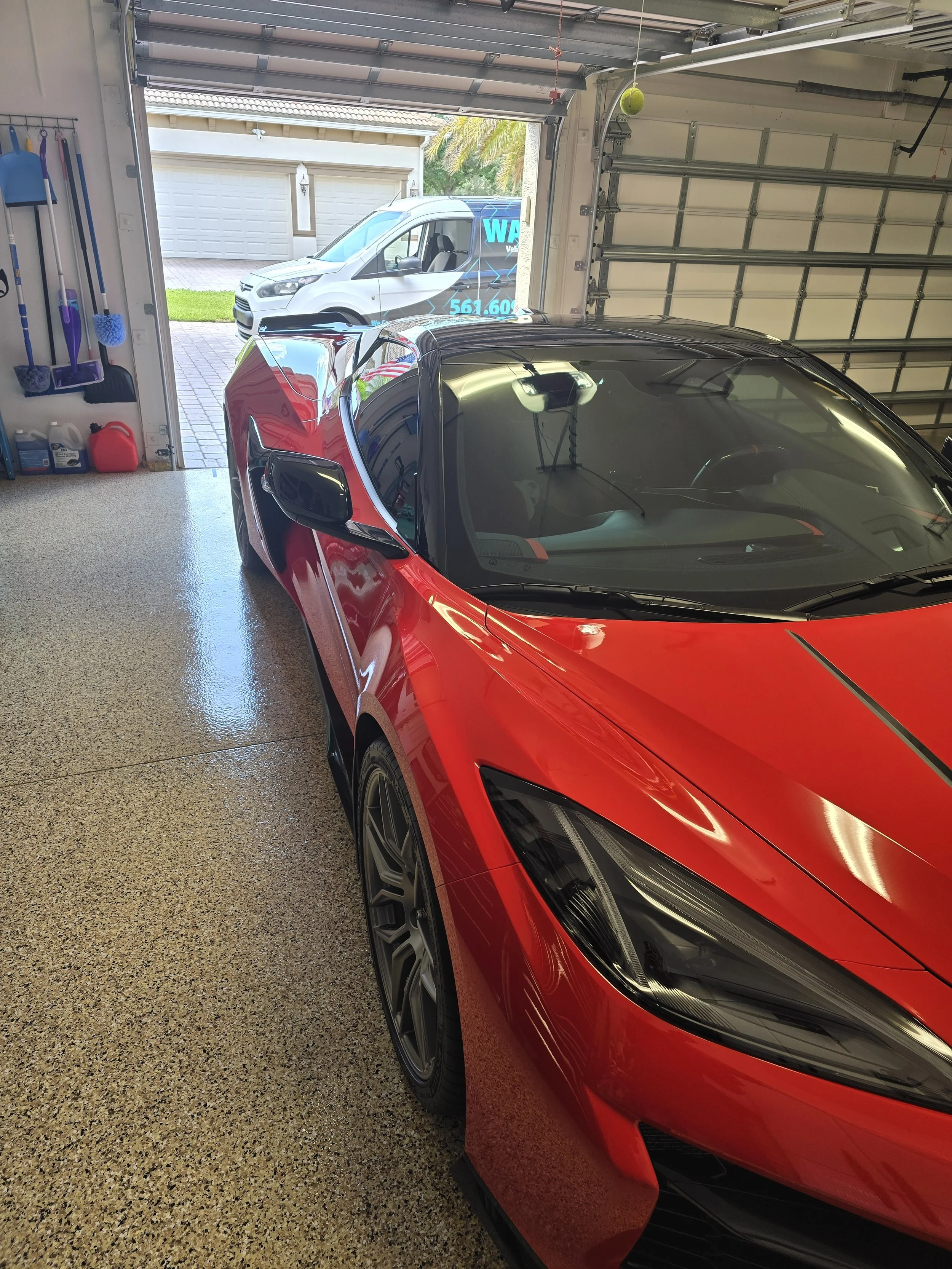 A red sports car parked inside a garage with an open garage door. Outside, a white van is visible on a driveway with a two-car garage in the background.