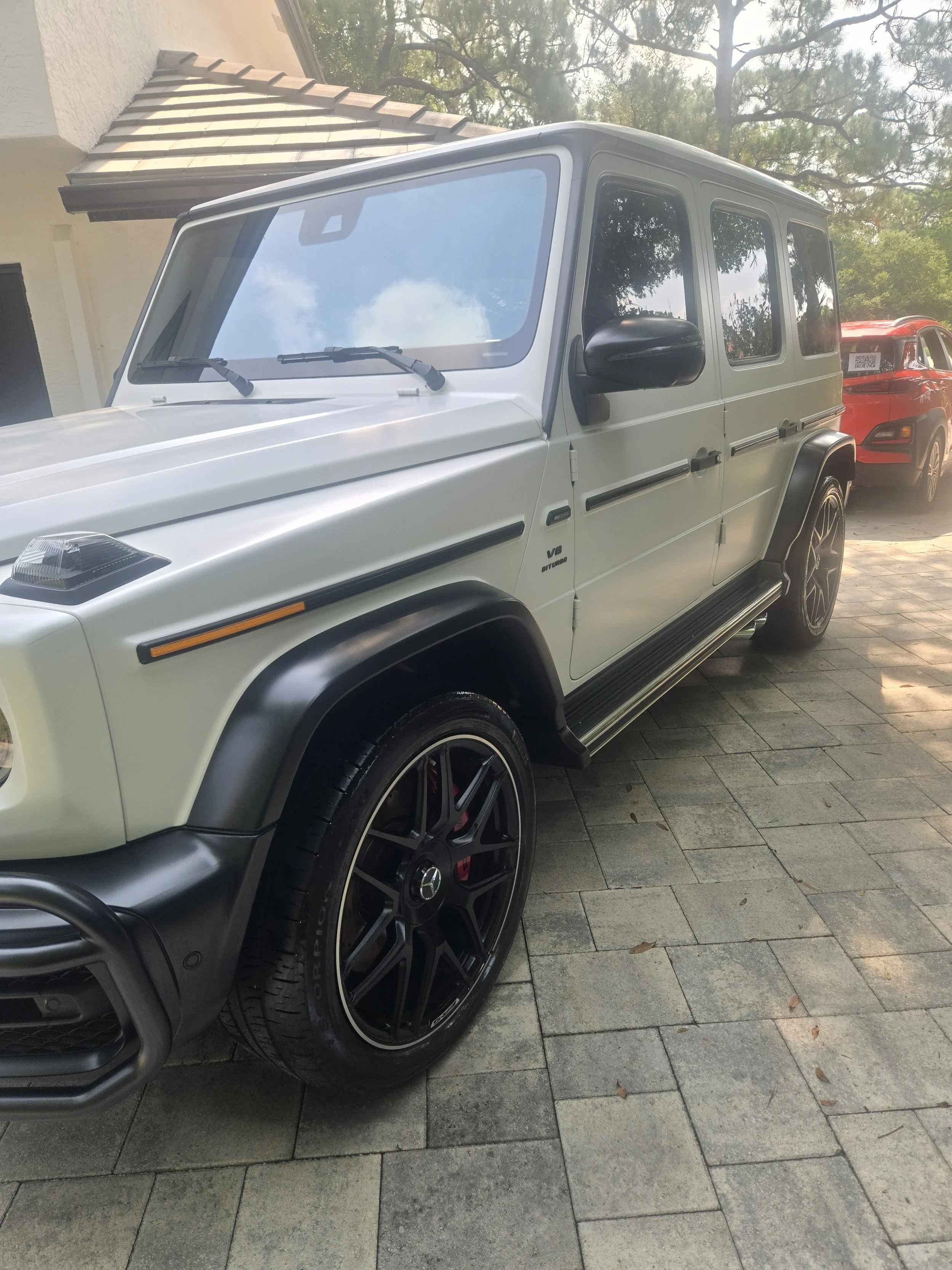 A white Mercedes-Benz G-Class SUV parked on a stone driveway next to a red vehicle, with a house's beige wall and roof in the background, trees overhead, and fallen leaves on the ground.