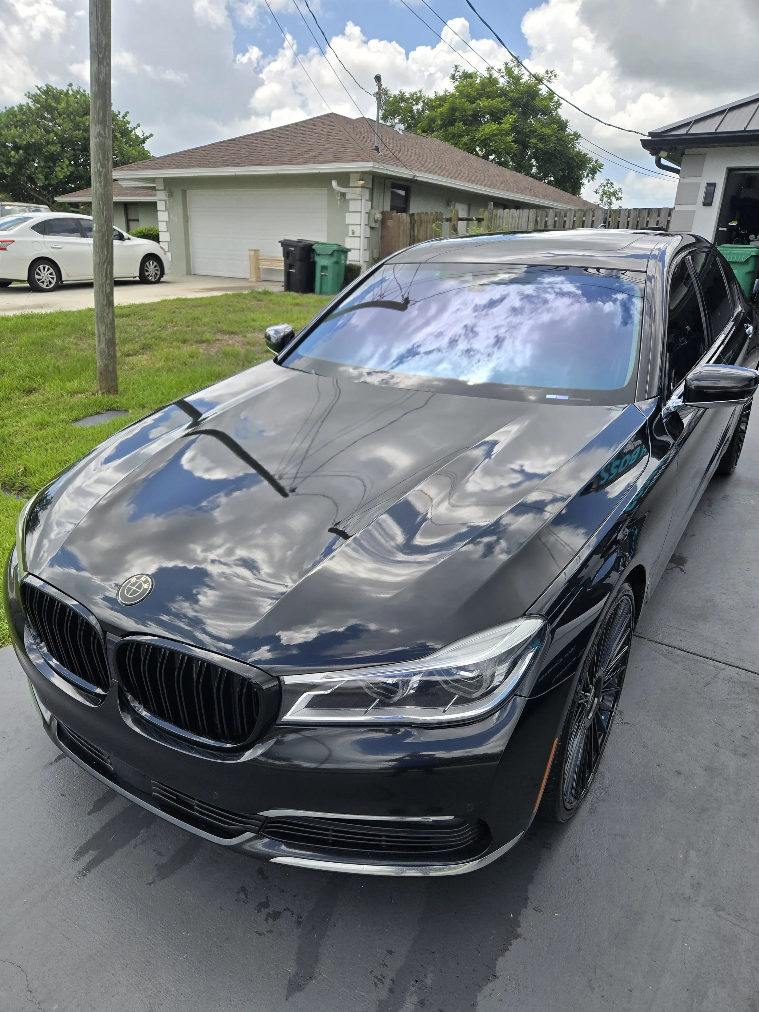 Black Mercedes-Benz car parked on driveway with reflection of clouds on windshield, suburban house in background with garage door and waste bins outside.