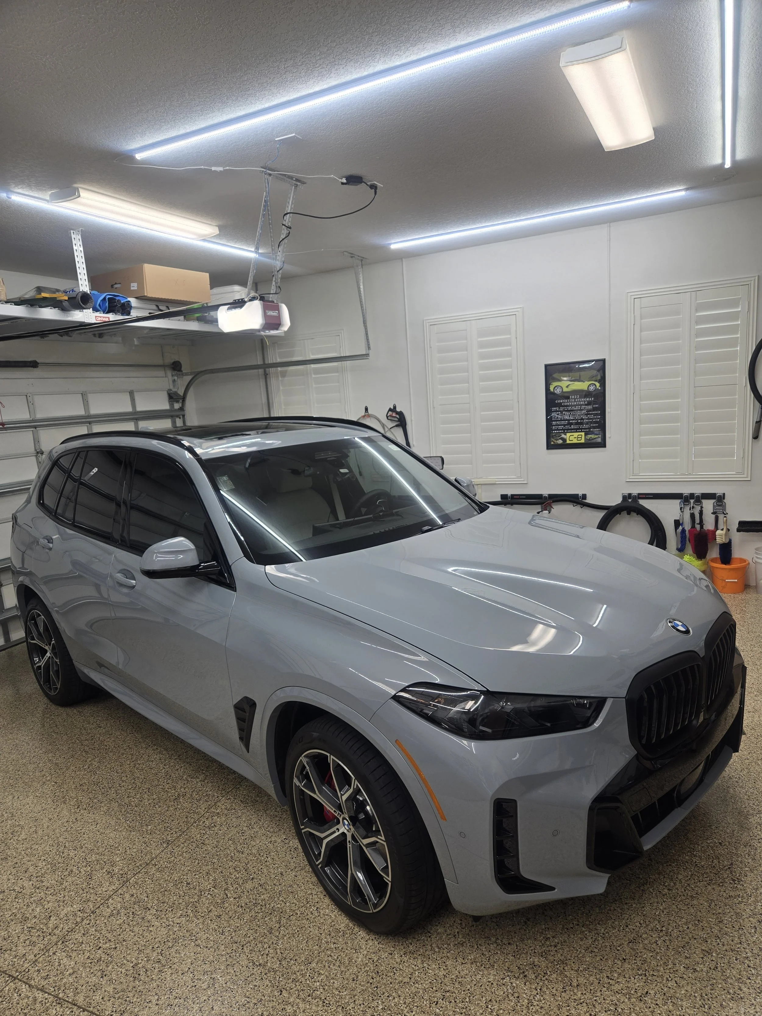 A gray BMW SUV parked inside a well-lit garage with fluorescent lighting, tools, and storage shelves visible on the walls.