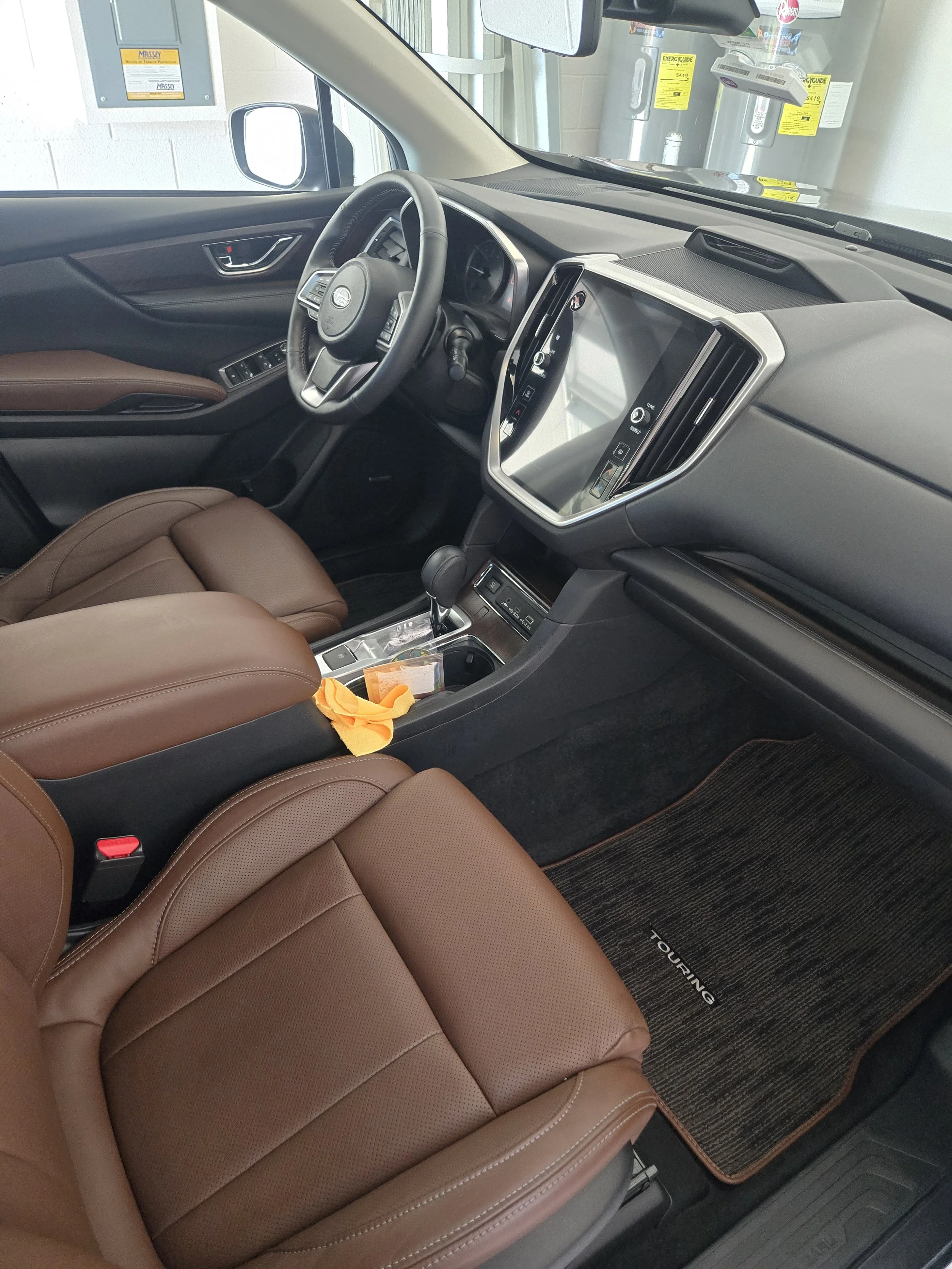 The interior of a vehicle showing a brown leather seat in the foreground and the dashboard with a touchscreen display, steering wheel, and gear shift in view. There are items in the center console including an orange cloth and a small plastic bag. Th