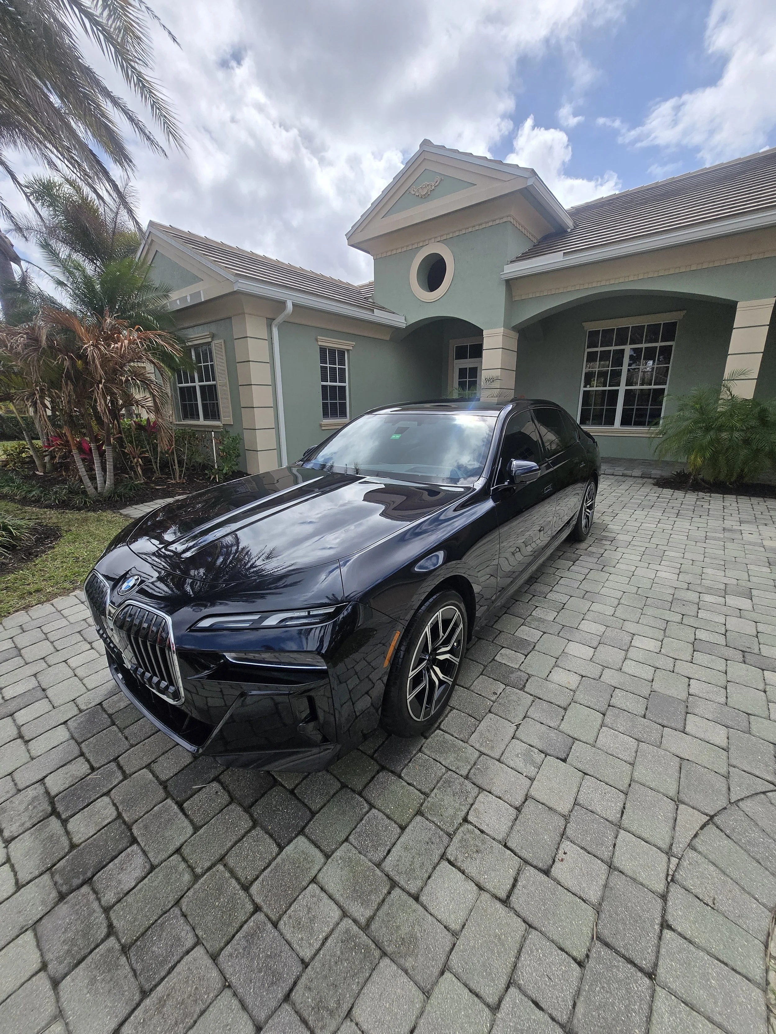A black BMW car parked in front of a light green house with palm trees and a cloudy sky.