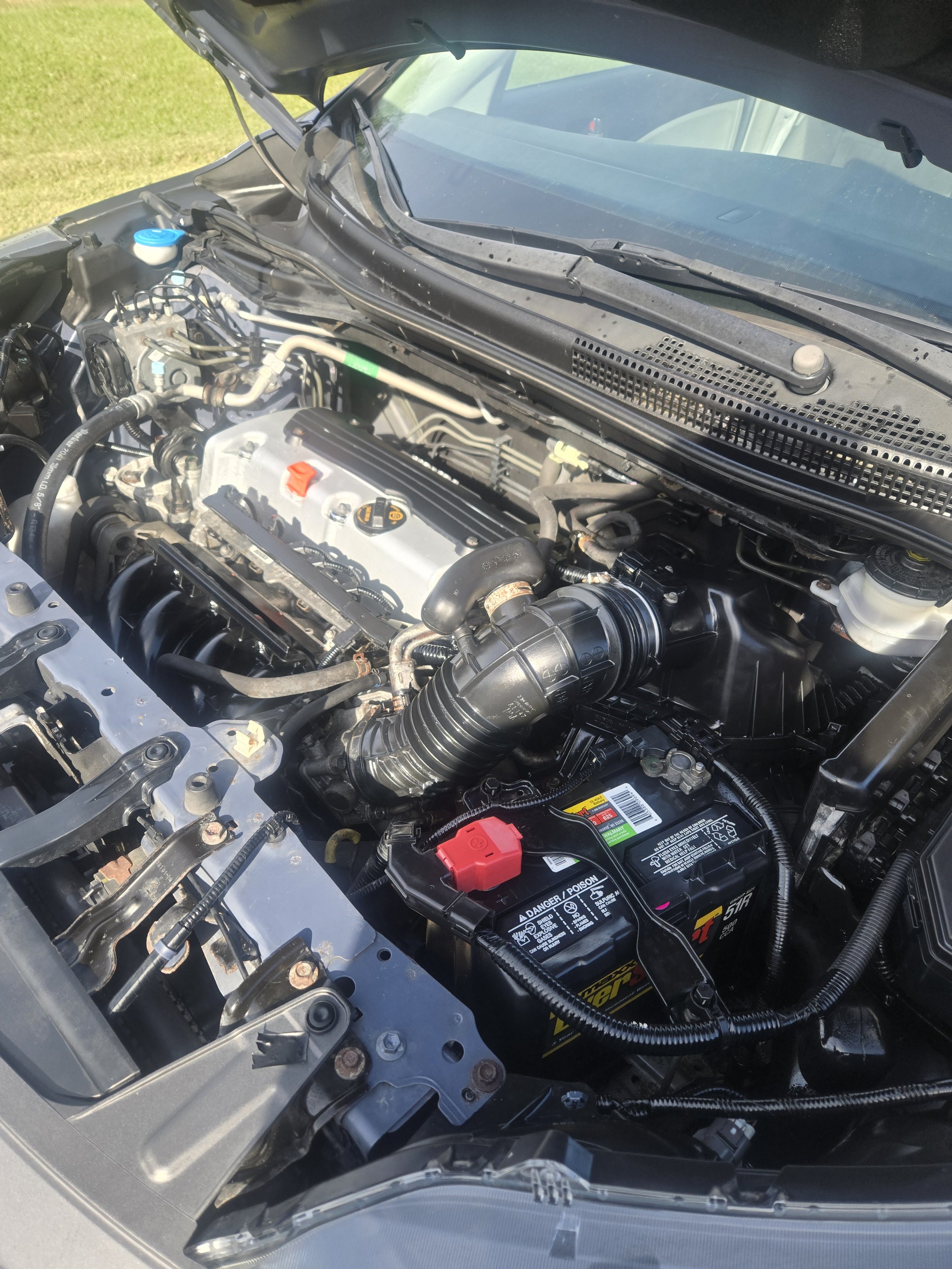 Close-up view of a car engine bay, showing various engine components including the battery, air intake, and engine block with valve covers. The hood is open and clipped to hold it up.