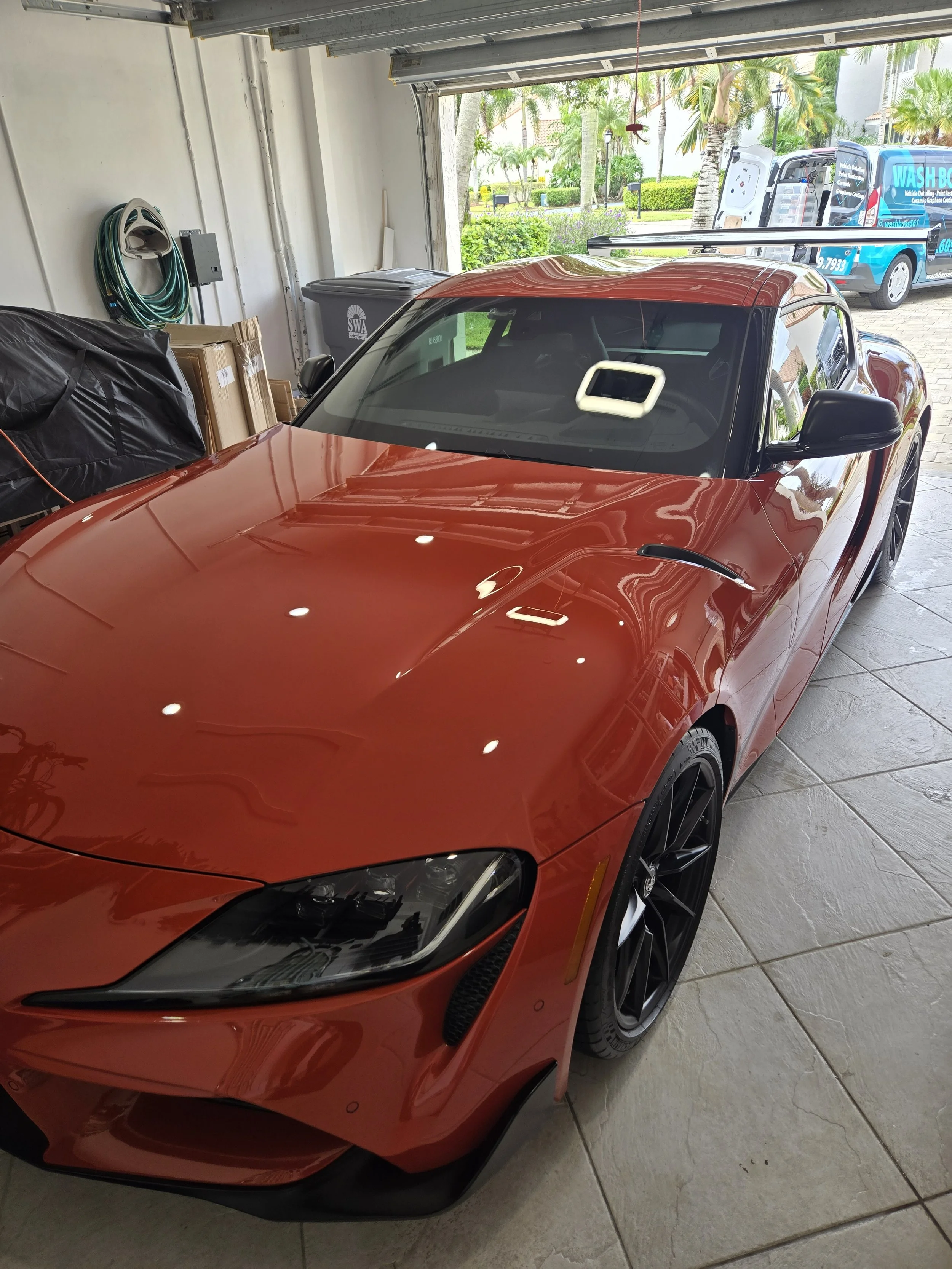 Red sports car parked inside a garage, with the garage door partially open revealing palm trees and parked vehicles outside.
