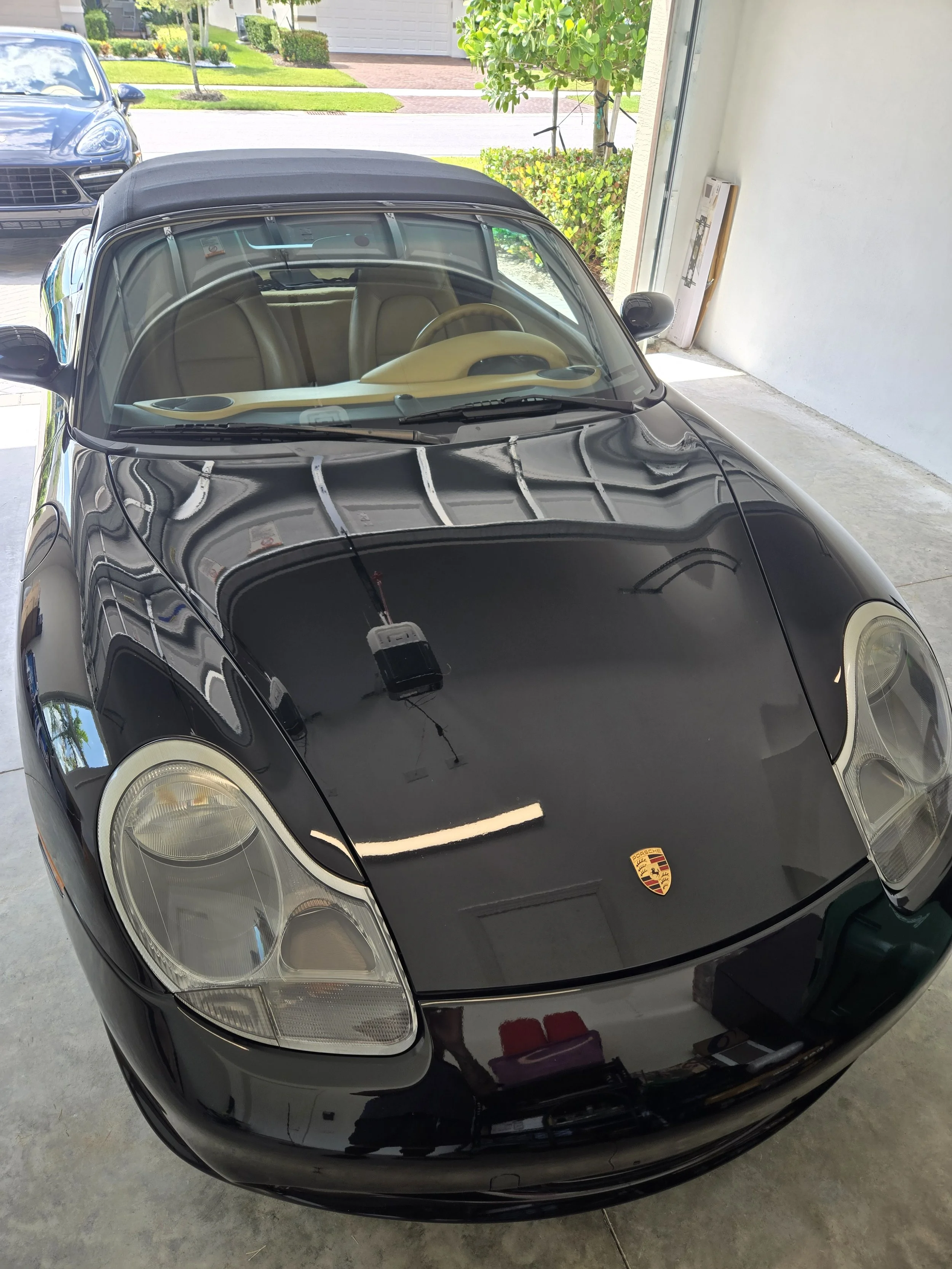 Black Porsche convertible with beige interior parked in a garage with a porch and green lawn visible outside.