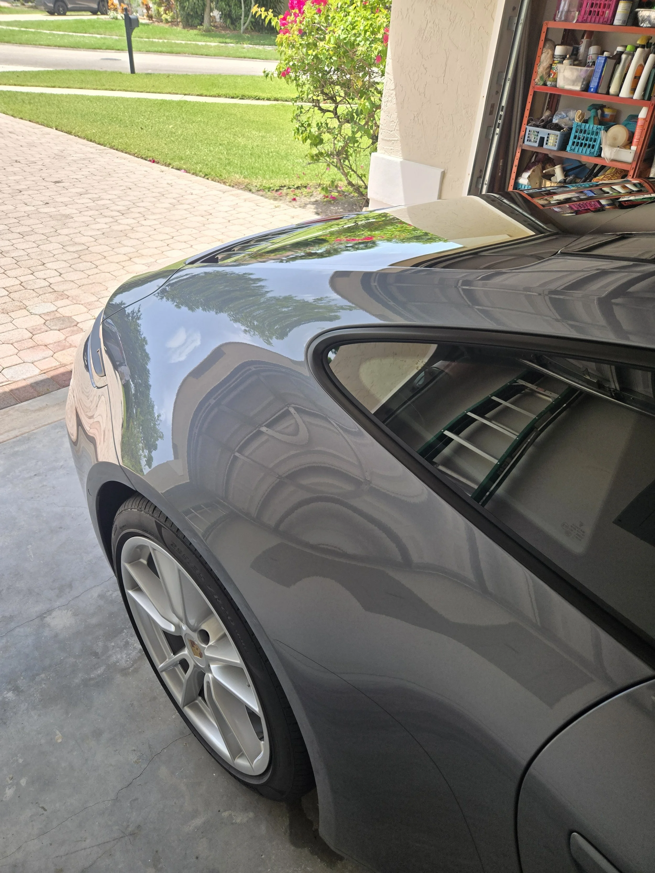 Partial view of a gray car parked in a driveway with a reflection of trees and the sky on its hood, next to a garage with shelves containing various bottles and tools.