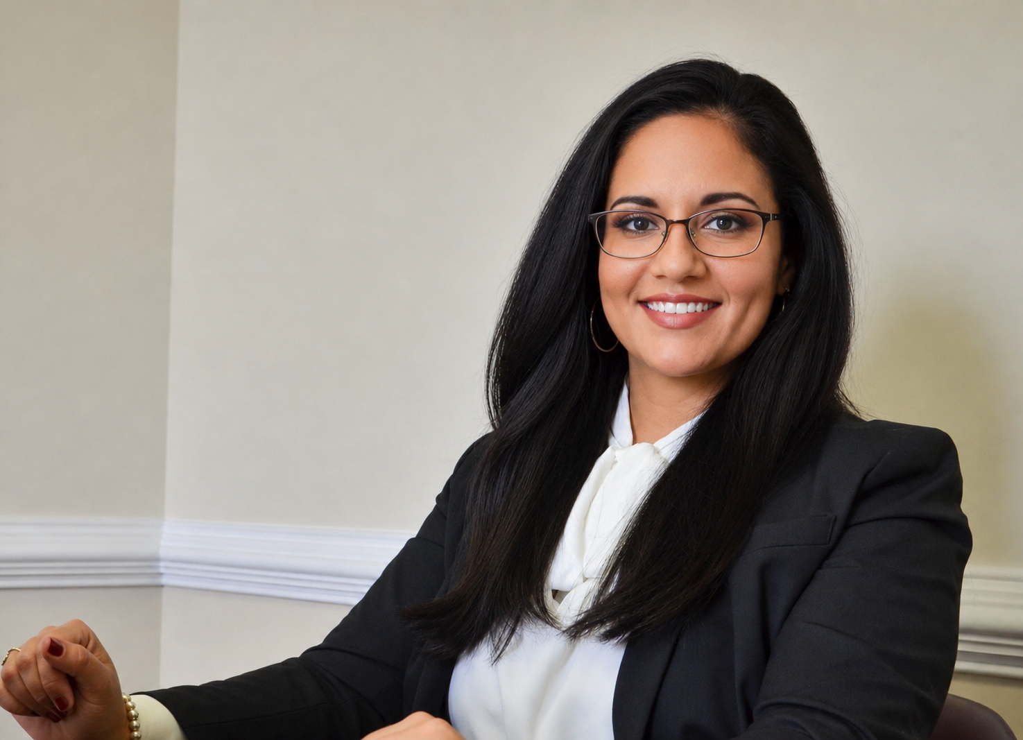 A professional woman with long dark hair, wearing glasses, a black blazer, and a white blouse, smiling confidently, sitting at a table in a neutral-colored room.