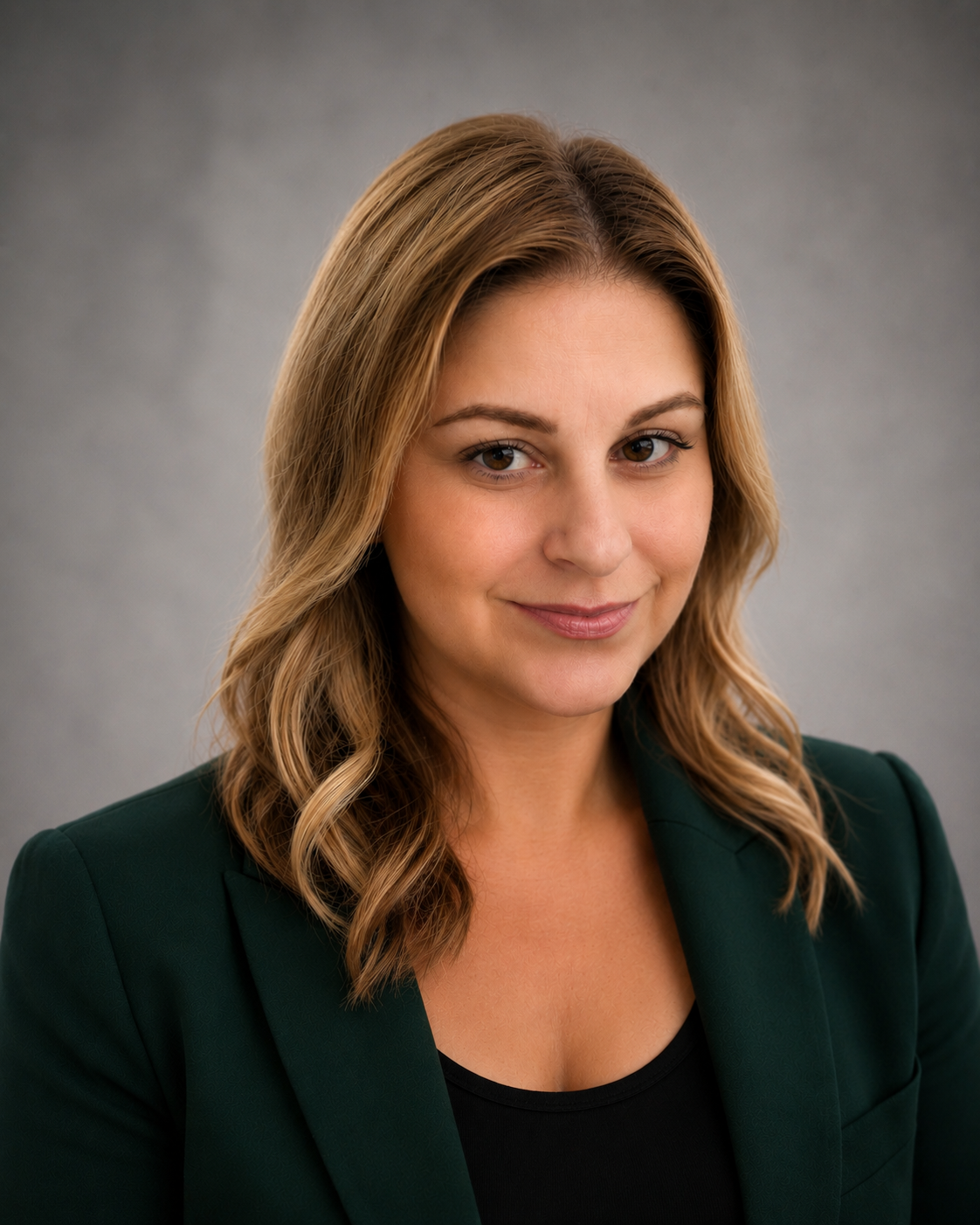 Portrait of a smiling woman with wavy, shoulder-length blonde hair, wearing a dark green blazer and black top, against a plain grey background.
