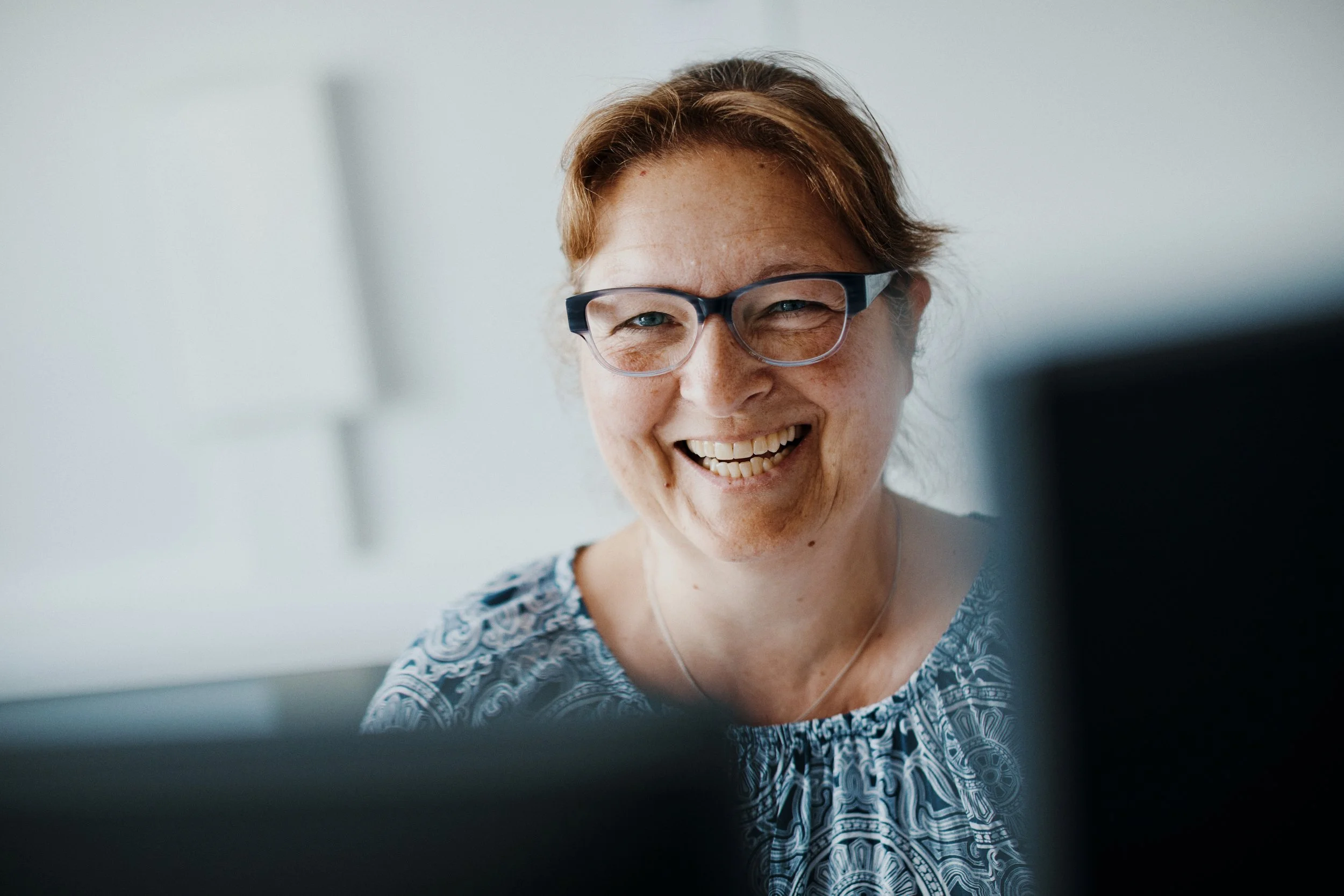 Lächelnde Frau mit Kurzhaarschnitt, Brille und blaus-weißem Muster-Shirt vor einem weißen Hintergrund, beim Arbeiten an einem Computer.