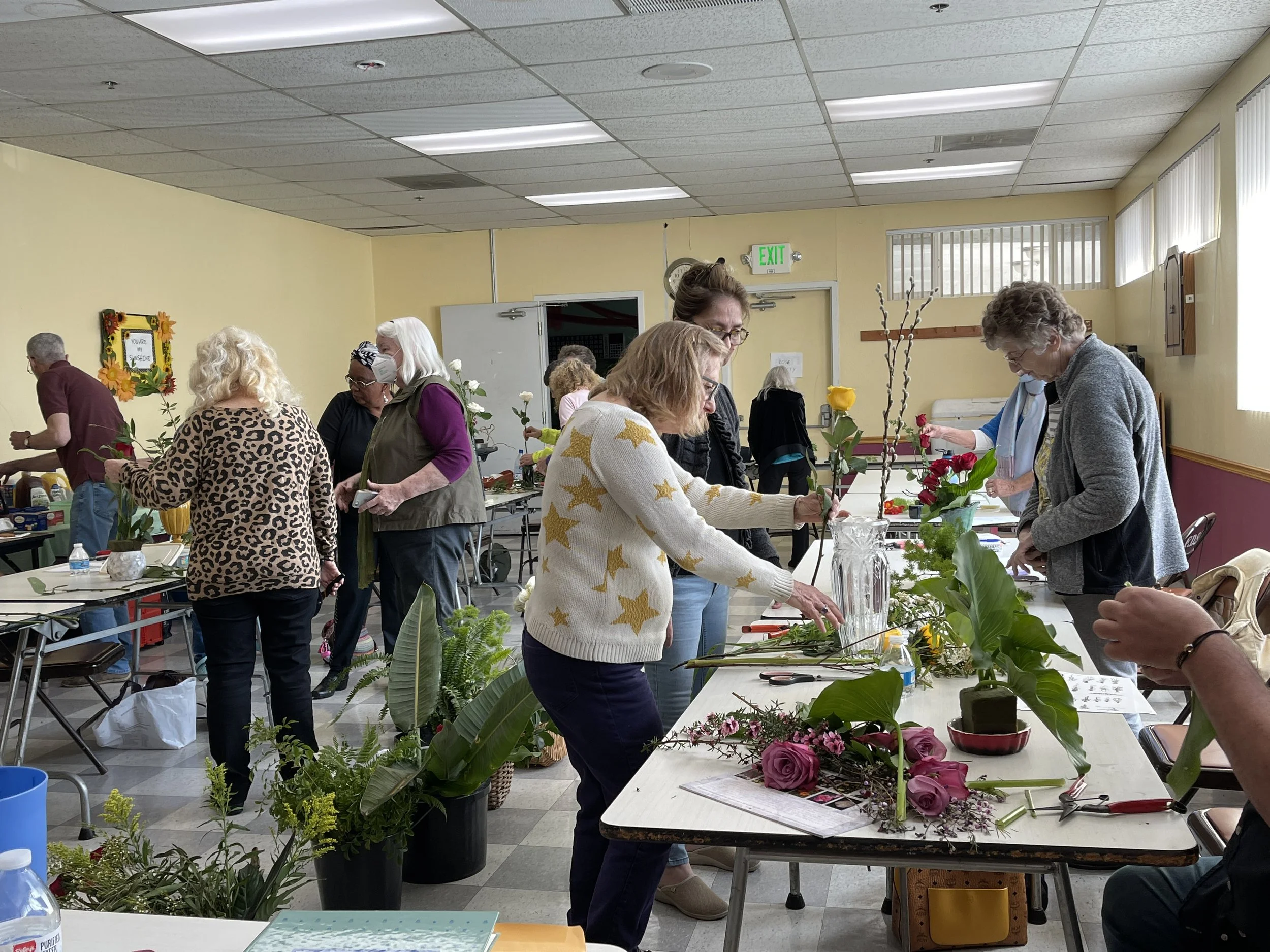 A group of people gathered in a room, working with flowers and plants at a floral arrangement workshop. They are standing by tables with various flowers, greenery, and tools, carefully arranging floral displays.
