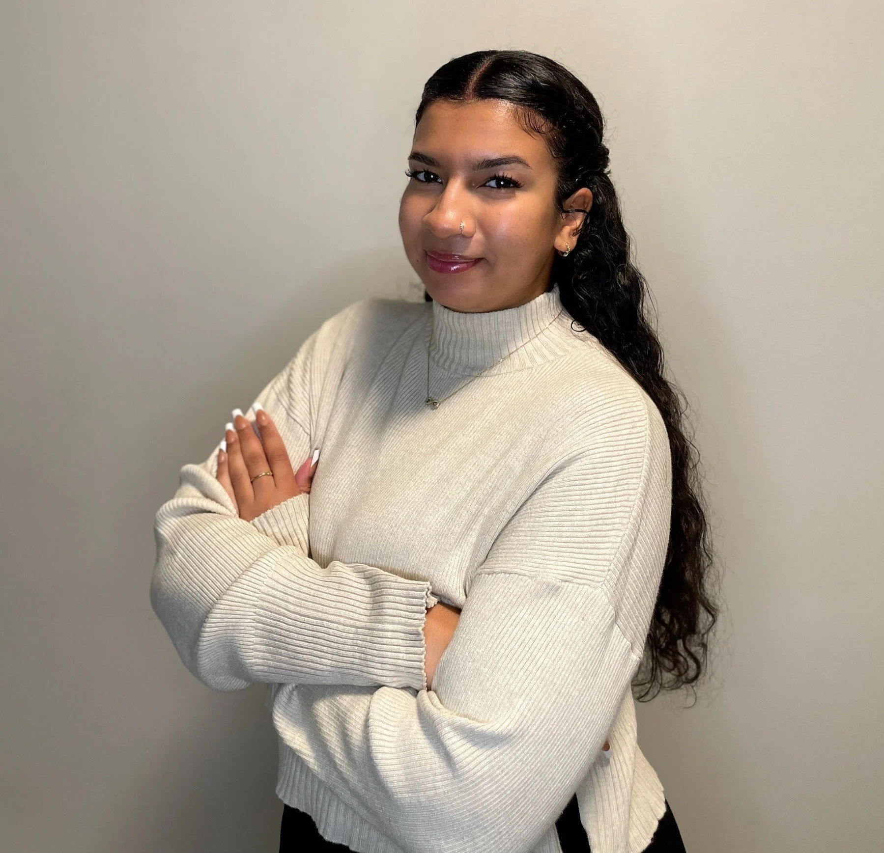 A woman with dark, curly hair and light brown skin, wearing a beige turtleneck sweater, looking at the camera with a slight smile and folding her arms.