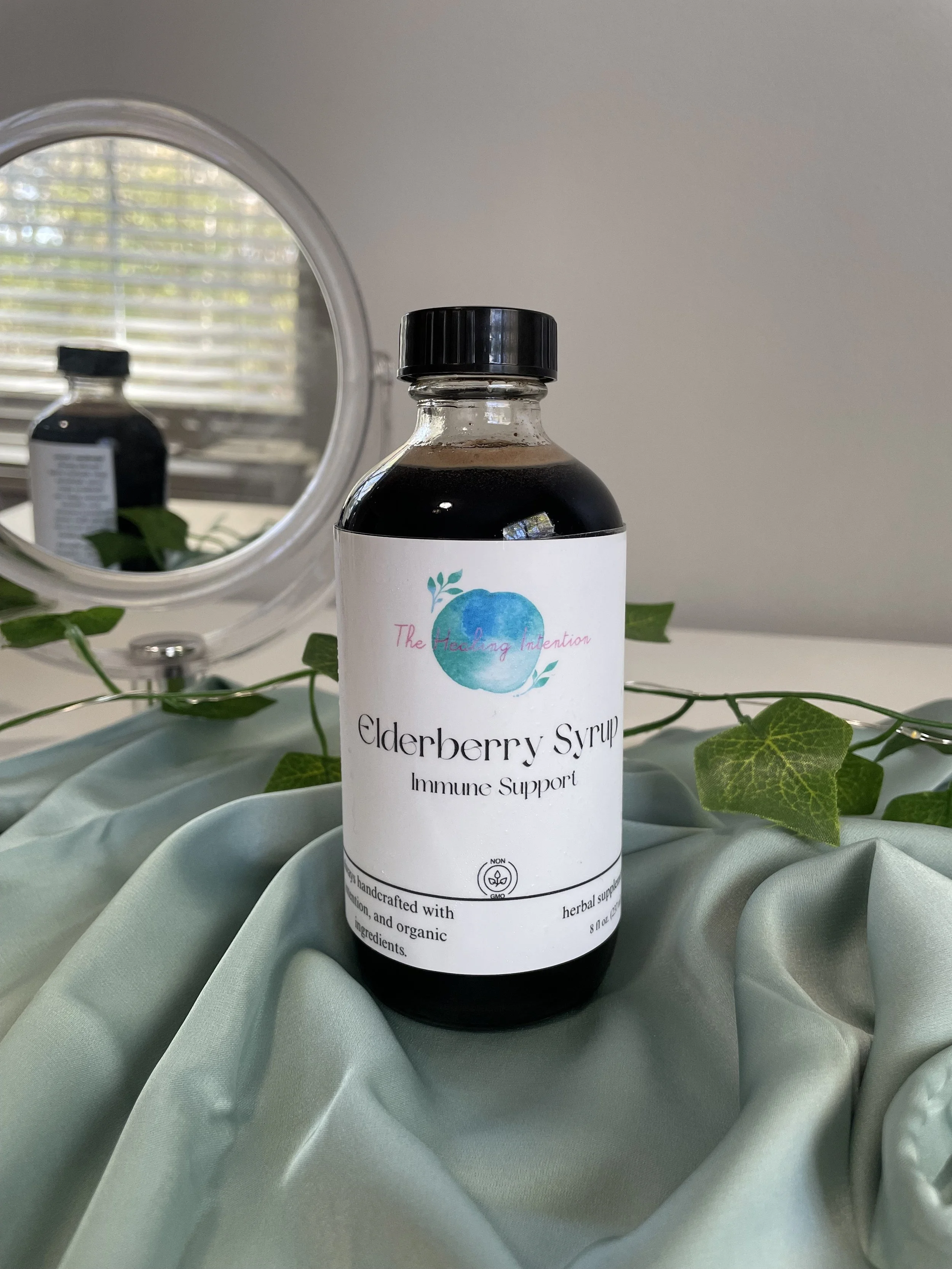 Bottle of elderberry syrup labeled 'The Healing Intention' with a logo featuring a blue fruit and green leaves, placed on light green fabric with green ivy leaves, and a mirror reflecting a window.