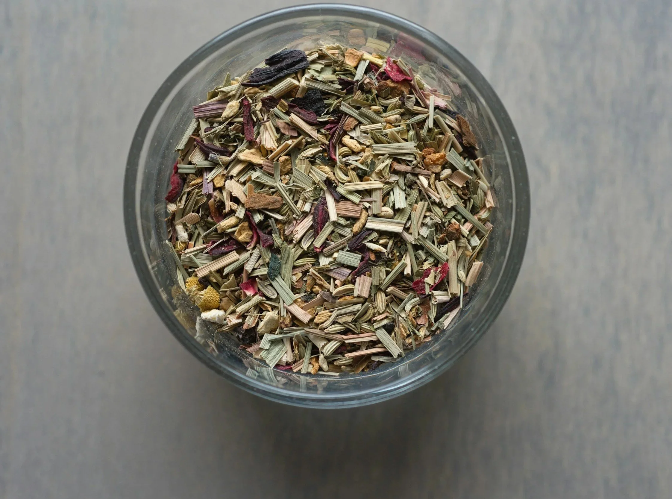 Top-down view of a clear glass bowl filled with dried herbal tea blend containing various herbs, flowers, and leaves.