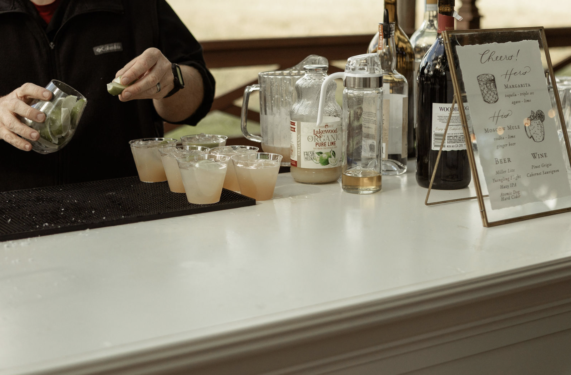Person preparing cocktails at a bar with various bottles, a pitcher of lime juice, and a menu board listing drinks and wine.