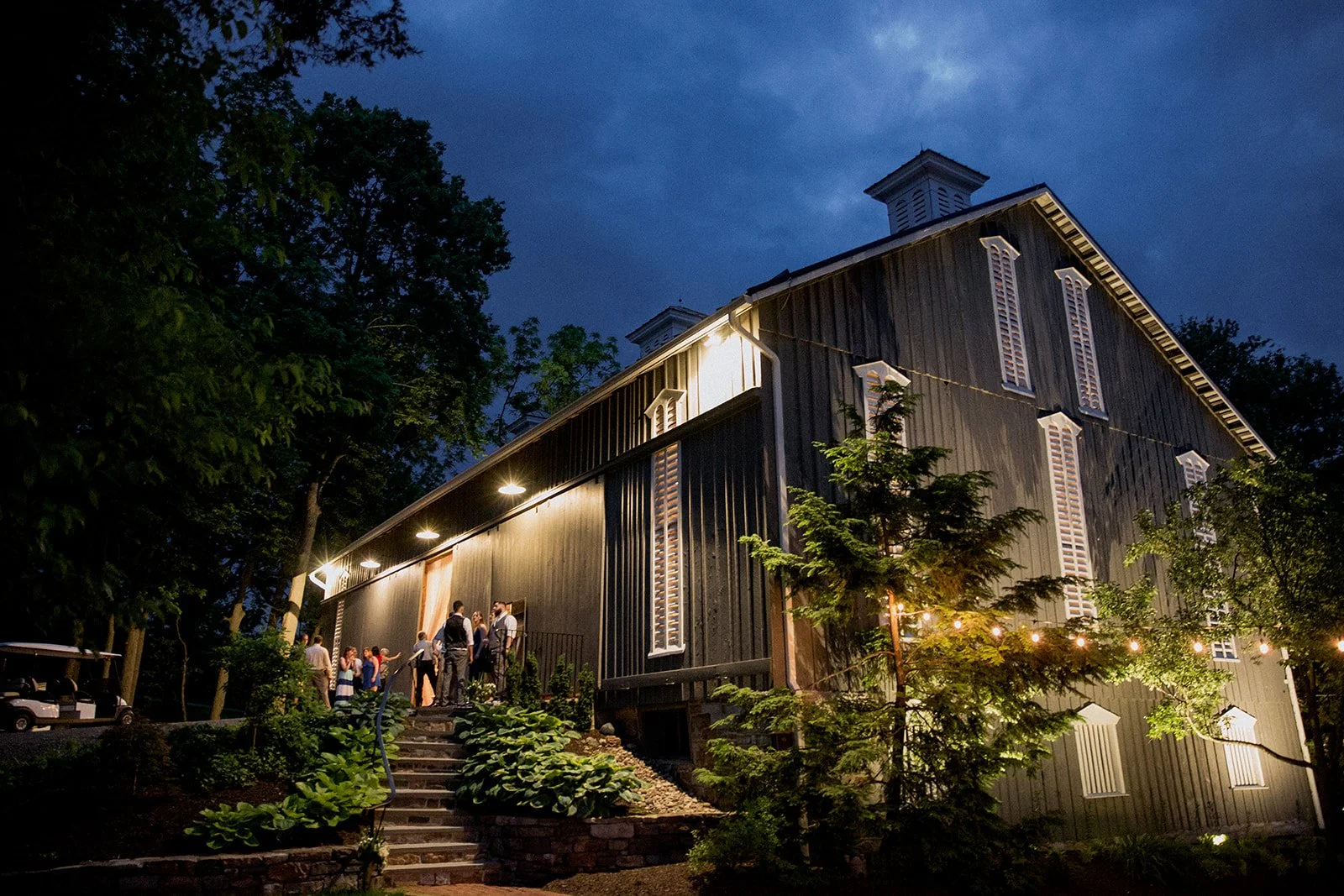 People standing on steps outside a dark wooden barn building illuminated at night, surrounded by trees with a cloudy evening sky in the background. This is One Barn Farm in Mifflinburg PA near Lewisburg PA.